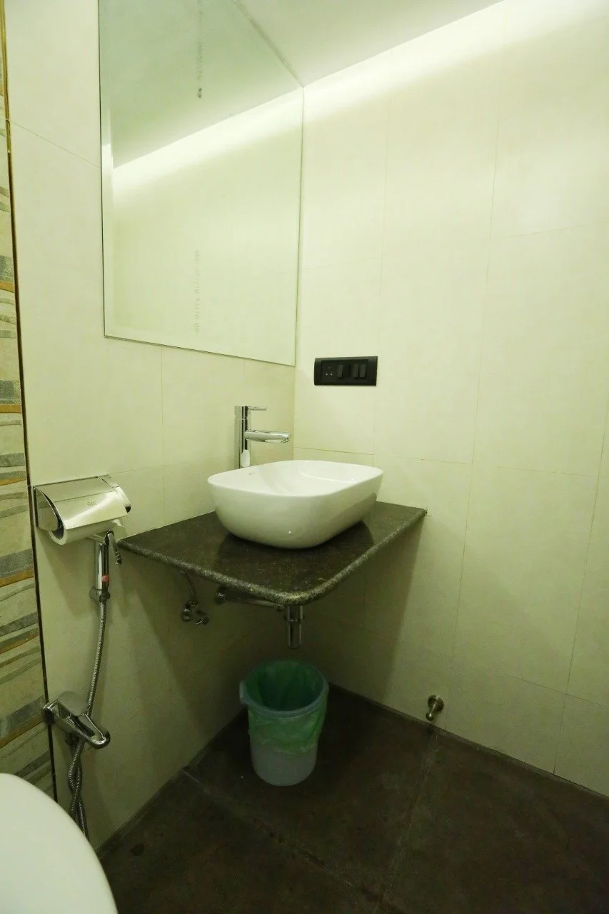 A bathroom with a sink on a dark granite countertop, a mirror on the wall, a waste bin underneath, a sleek faucet, and a wall-mounted toilet paper holder with a bidet spray, all with minimalistic decor.