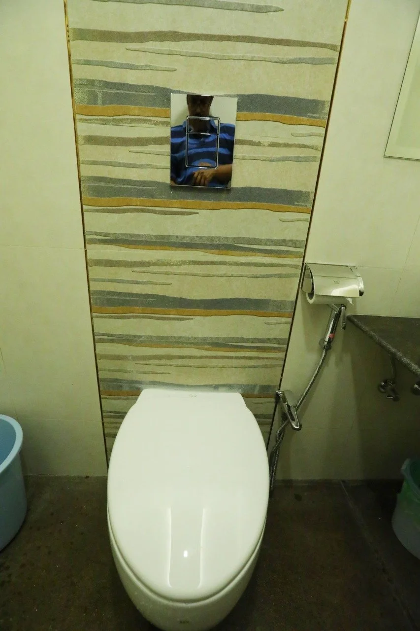 Image of a modern toilet in a bathroom, with a textured wall behind and a mirror above, reflecting a person taking the photo.