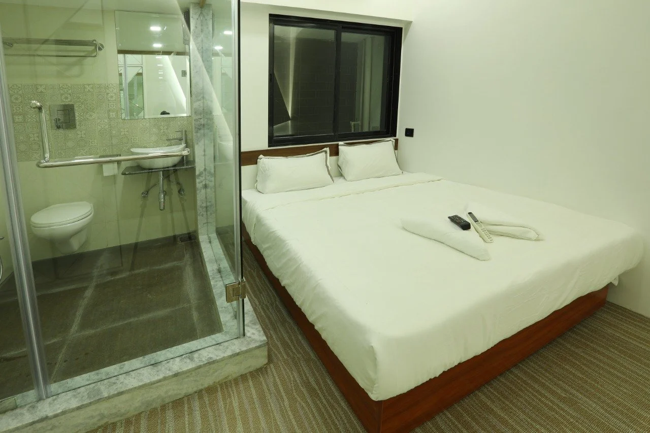 A hotel room featuring a neatly made bed with white linens, a window behind the bed, and a built-in bathroom with a glass wall, toilet, and a sink.