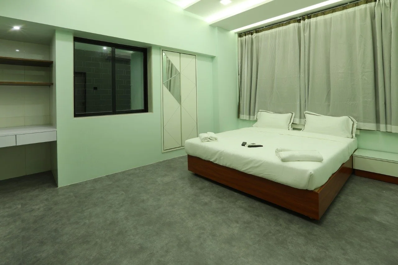A clean hotel room with a bed, folded towels, remote controls, a window with curtains, and a small shelf.