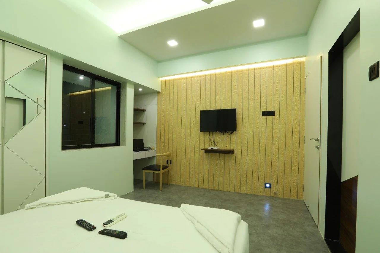 Modern hotel room with a wall-mounted TV, a desk with a chair, a window, and a bed with remote controls on it.