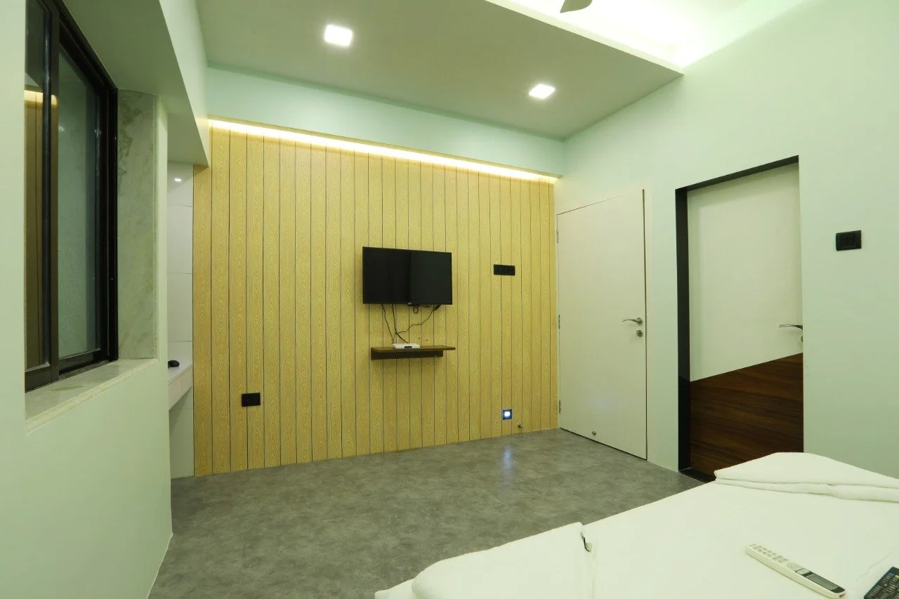 Modern bedroom with a mounted TV on a yellow wooden accent wall, a bed with white linens and remote controls, a window with a sliding panel, and a door leading to another room with wood flooring.