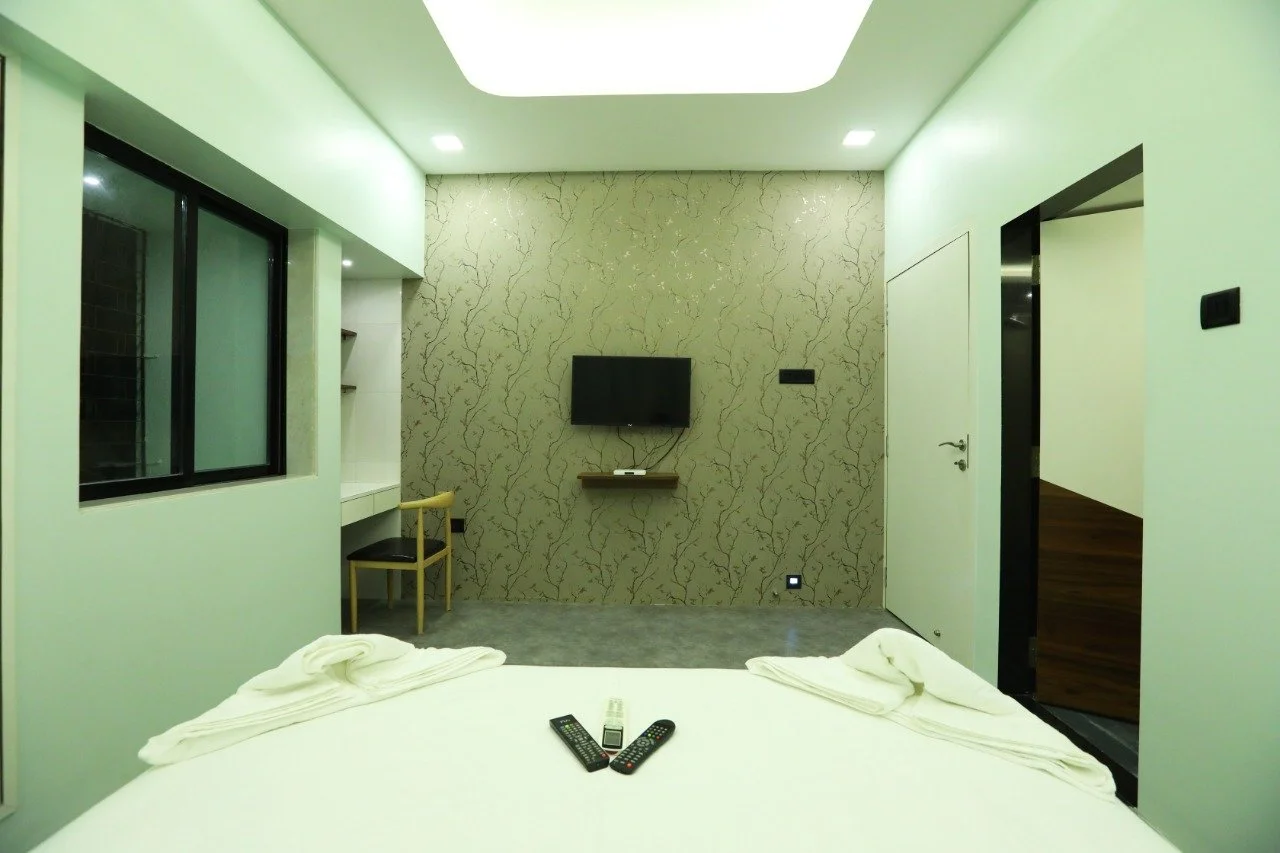 Hotel room with a bed, TV, window, desk, chair, and door, with remote controls on the bed.