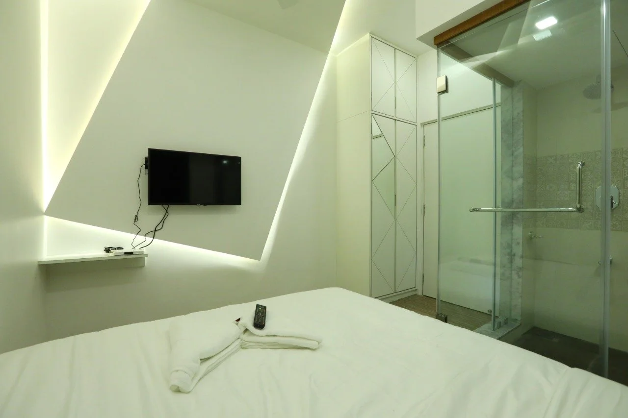A modern, minimalistic hotel room with a bed, a wall-mounted television, and a glass-enclosed bathroom with a walk-in shower.
