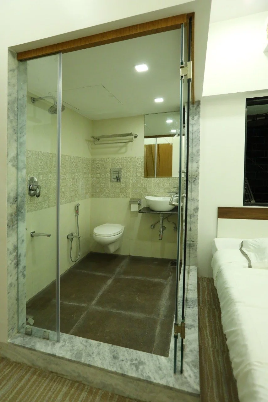 A modern hotel room featuring an open bathroom layout with a walk-in shower, toilet, and a small sink, visible from the main bedroom area with a bed and striped carpet.