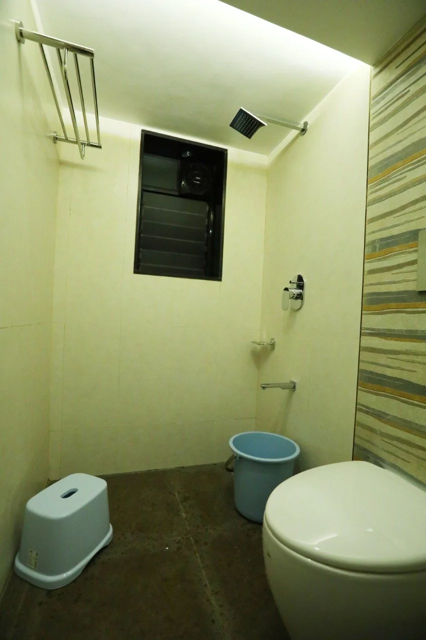 A small bathroom with a modern toilet, small blue bucket, window, handrail, soap holder, and a shower head on the wall.