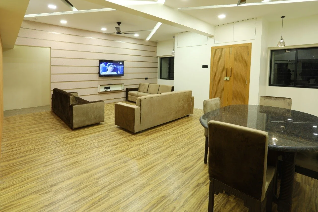Living room with beige sofas, mounted TV, wooden flooring, round dining table with chairs, and windows.