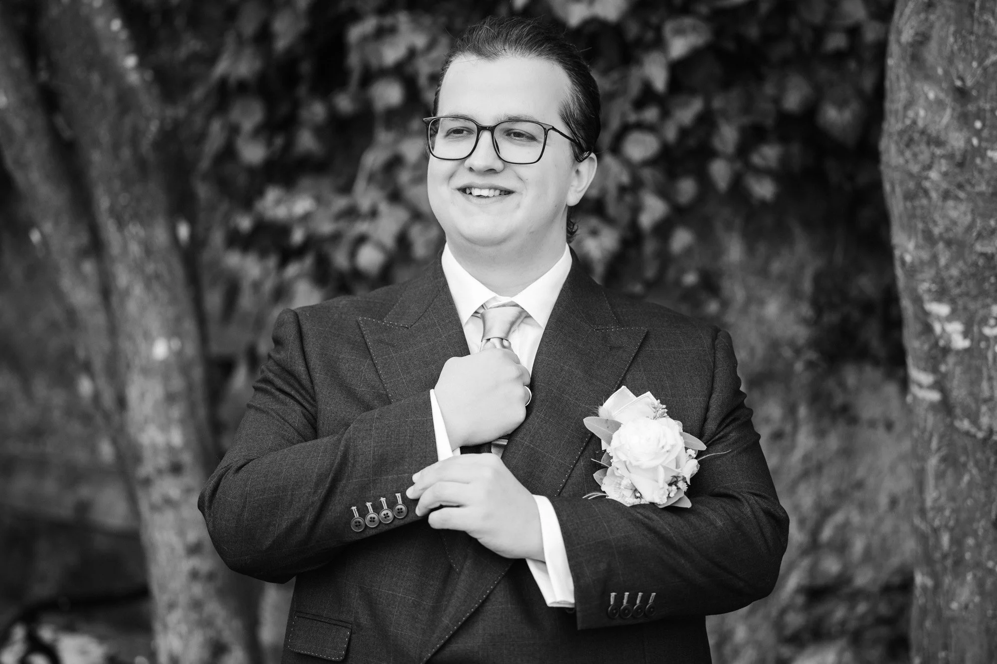 Schwarz-Weiß-Foto eines Mannes in Anzug mit Krawatte und Brille, der ein Lächeln zeigt, vor Baumstämmen und Blättern im Hintergrund.