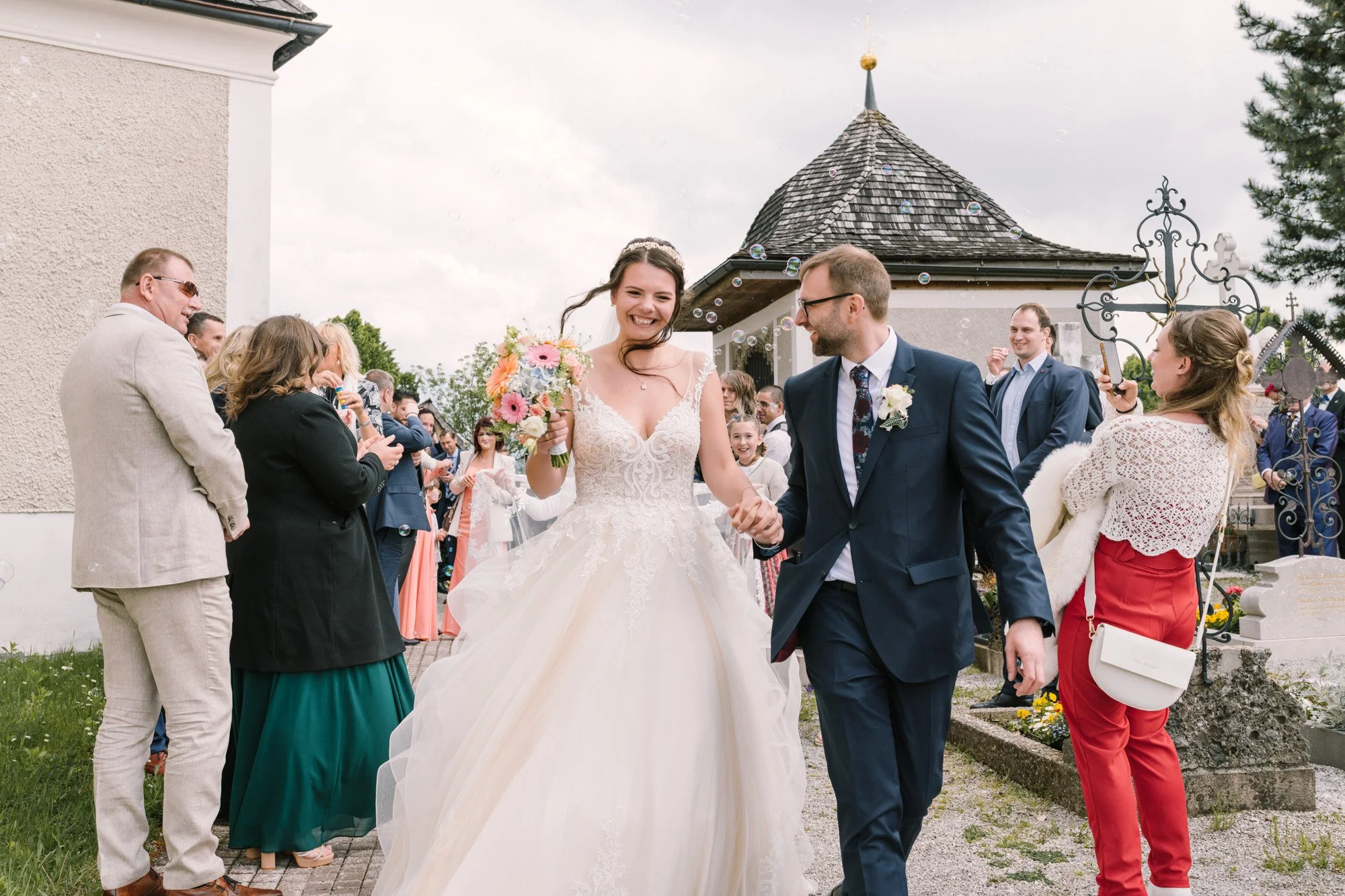 Braut und Bräutigam gehen bei ihrer Hochzeit im Freien Hand in Hand, umgeben von lachenden Gästen. Die Braut trägt ein weißes Hochzeitskleid mit Blumenstrauß, der Bräutigam trägt einen dunklen Anzug. Es gibt Seifenblasen im Himmel und eine historische Kapelle im Hintergrund.
