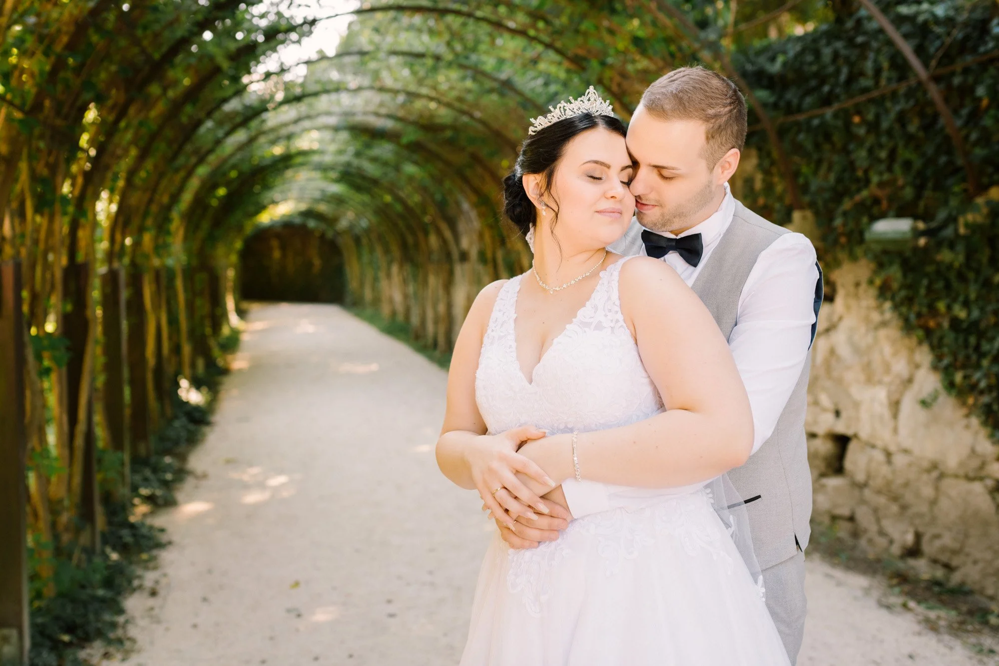 Ein Brautpaar steht eng beieinander in einem Tunnel aus Bäumen, beide in Hochzeitskleidung, umgeben von einem natürlichen, grünen Hintergrund.