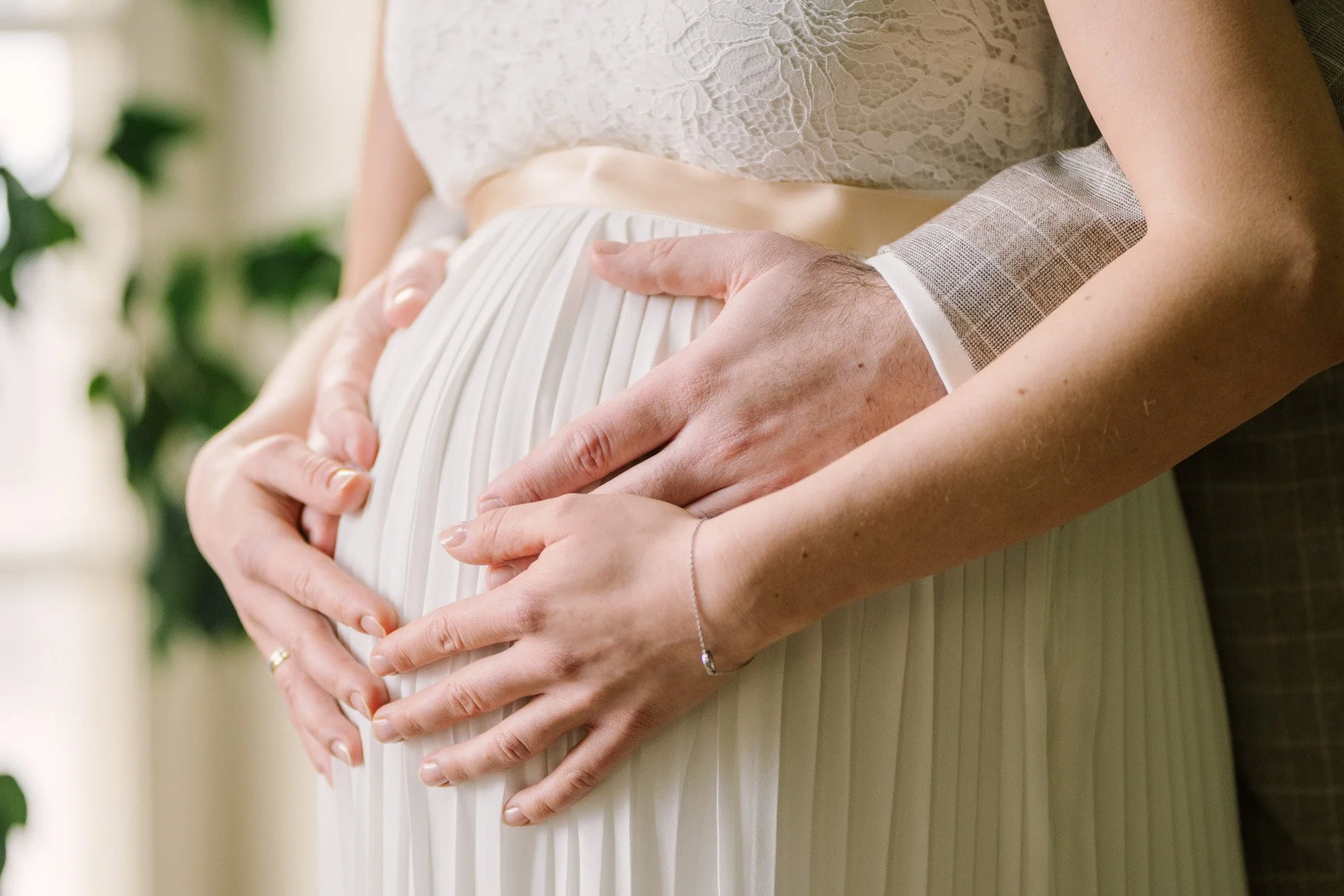 Nahaufnahme der Hände eines zukünftigen Elternteils, die den Babybauch einer schwangeren Frau halten, während sie ein elegantes weißes Kleid trägt.