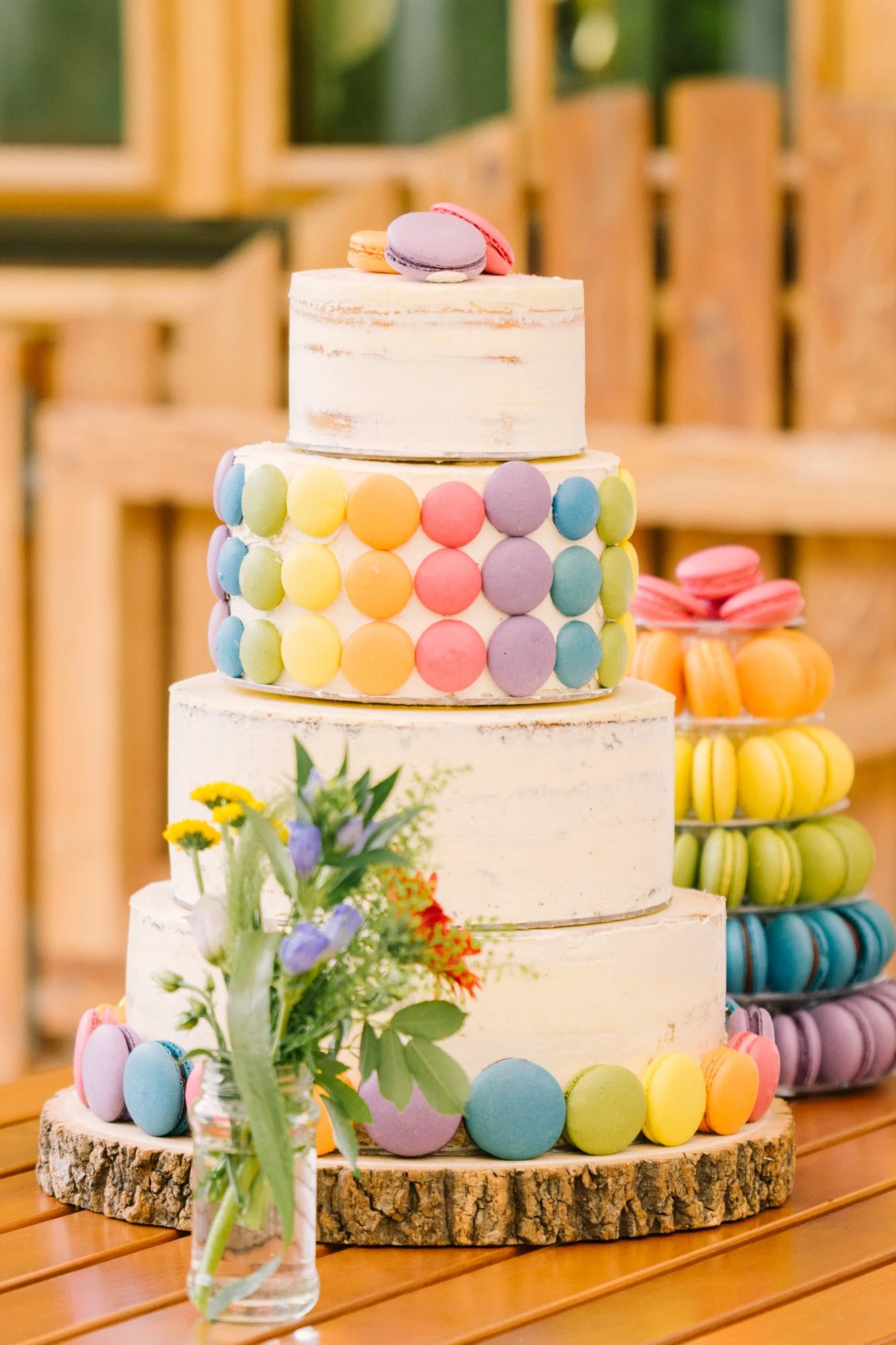 Mehrstöckiger Kuchen mit bunten Macarons und Dekoration aus Blumen auf Holzplatte, farbige Macarons im Hintergrund.