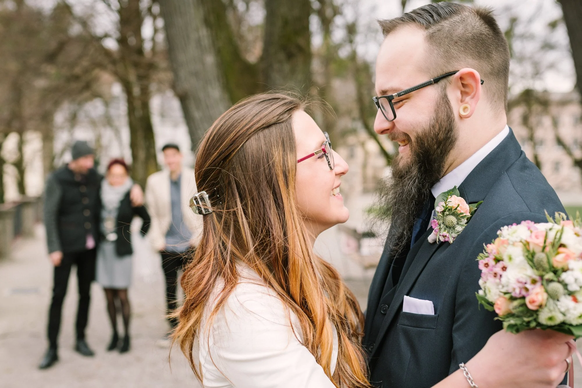 Ein glückliches Paar bei einer Hochzeit umarmt sich im Freien, im Hintergrund einige Freunde, Bäume und eine Parklandschaft.