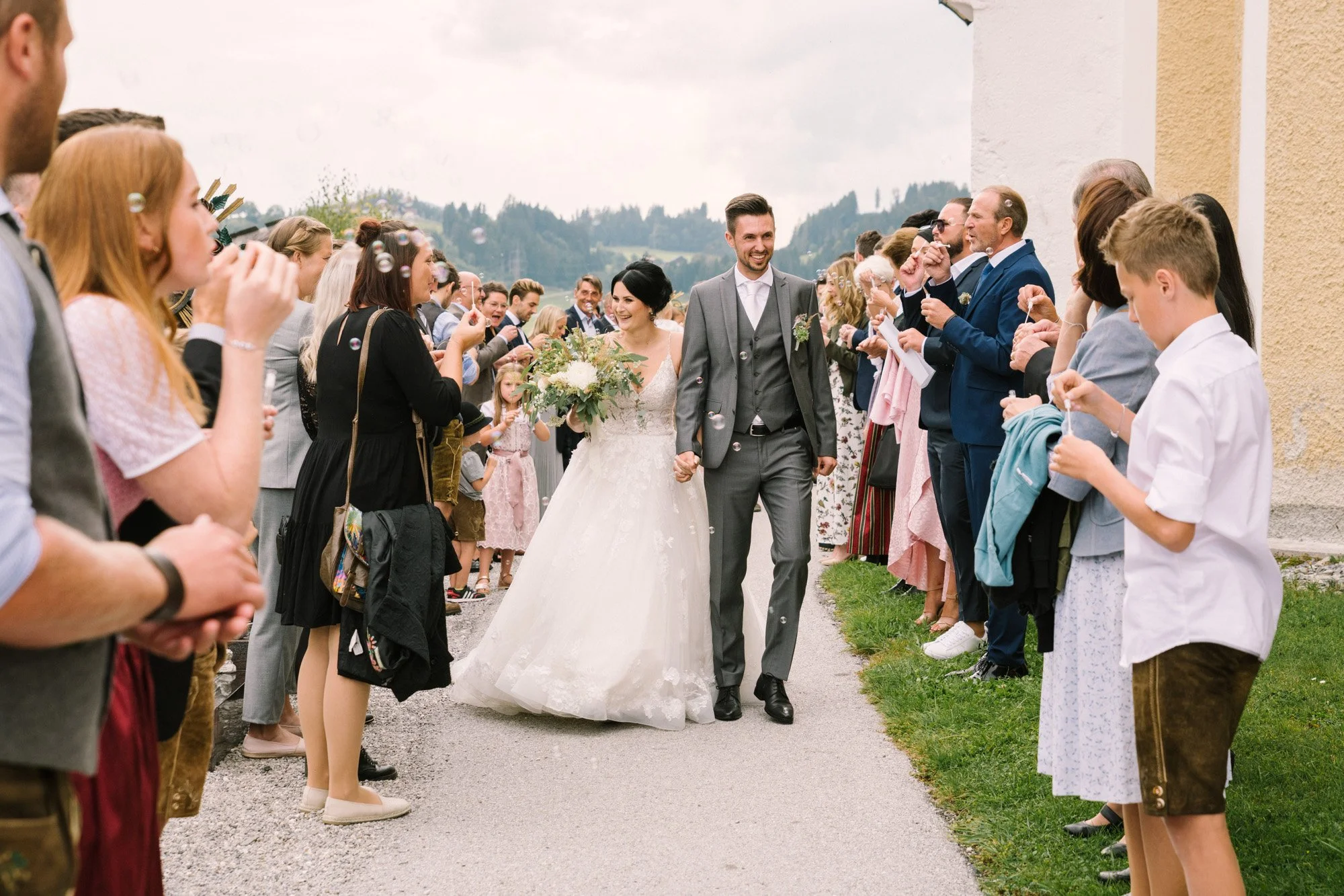 Ein Brautpaar läuft lachend Hand in Hand durch einen von Gästen eingerahmten Weg bei einer Hochzeit im Freien, umgeben von Freunden und Familie, im Hintergrund Berge und eine bewölkte Himmel.