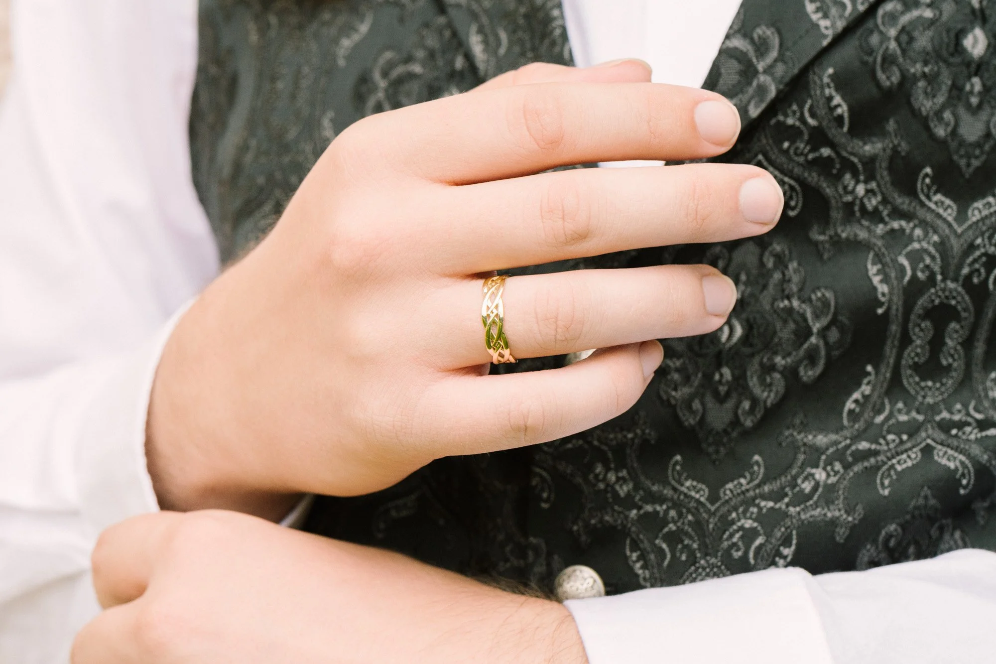 Nahaufnahme einer Hand mit einem goldenen Ring auf dem Ringfinger, die eine andere Person umarmt, im Hintergrund dunkle, paisley-verzierte Kleidung.