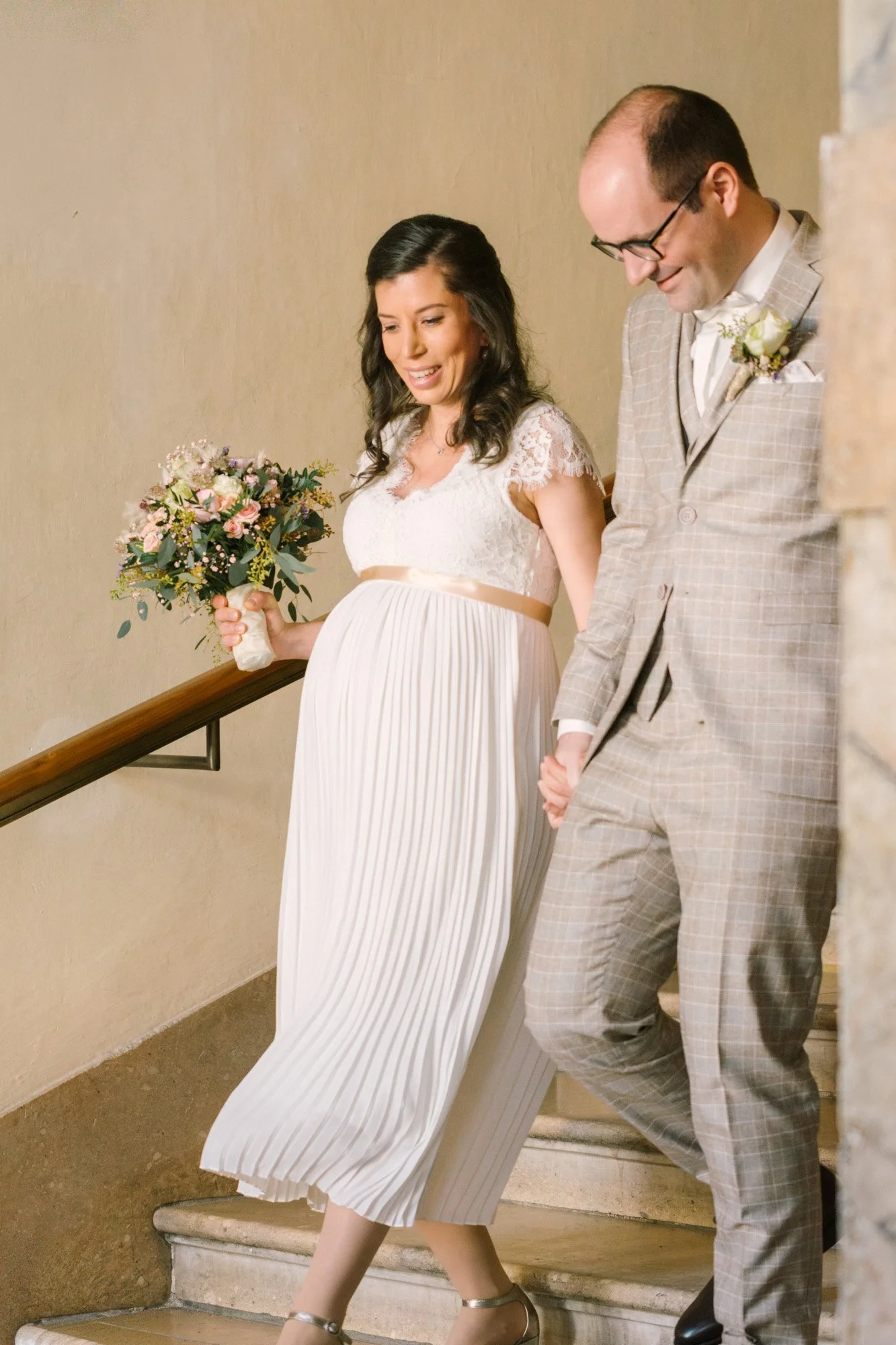Ein paar, das auf einer Treppe geht, bei einer Hochzeit. Sie trägt ein langes, weißes Kleid mit Plissees und hält einen Blumenstrauß, er trägt einen grauen Anzug mit Karo-Print, beide lächeln und sind im Moment der Zeremonie.