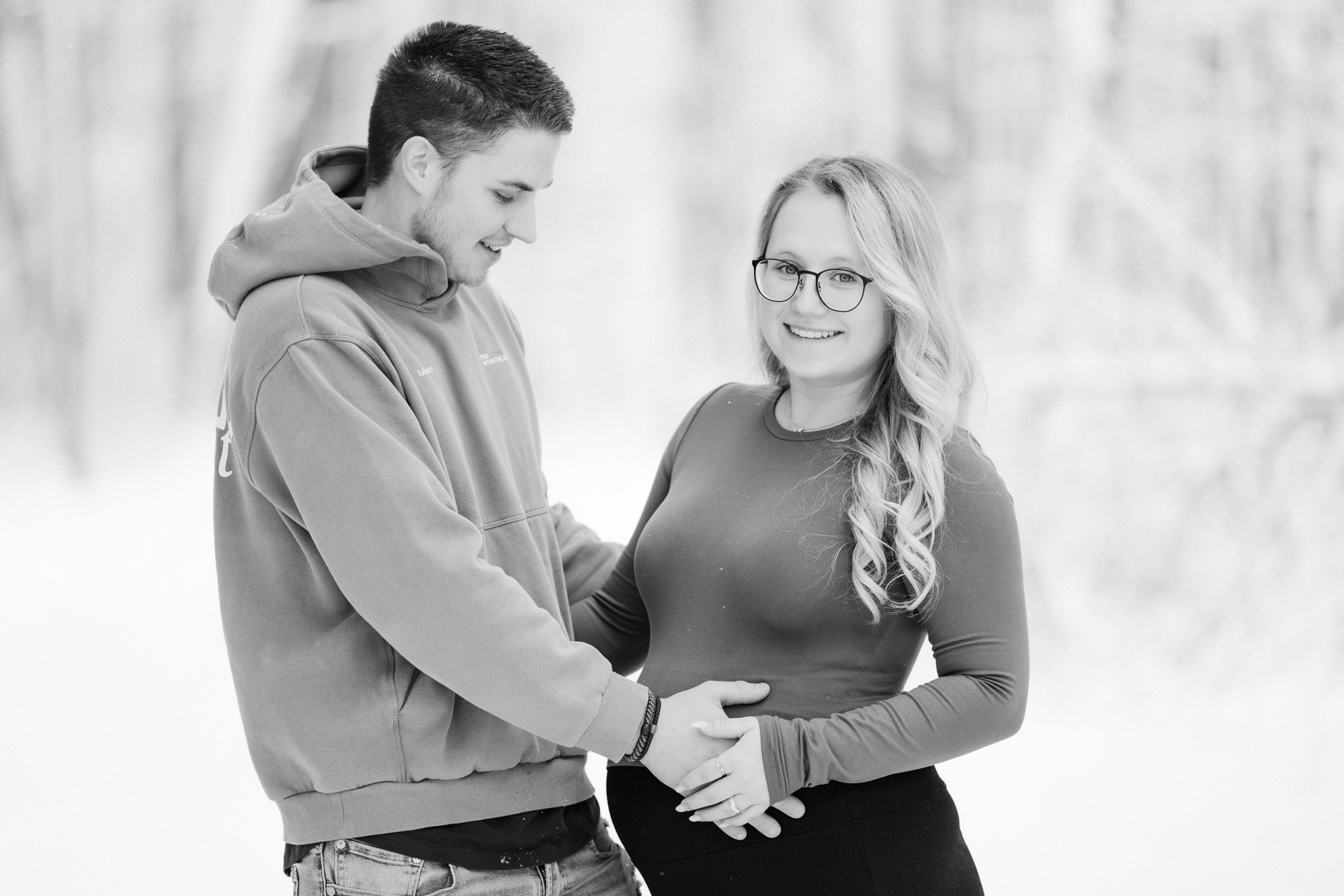 Ein lachendes stehendes Paar, ein Mann und eine schwangere Frau, die Hand auf den Bauch der Frau legen, im Freien im Winter.