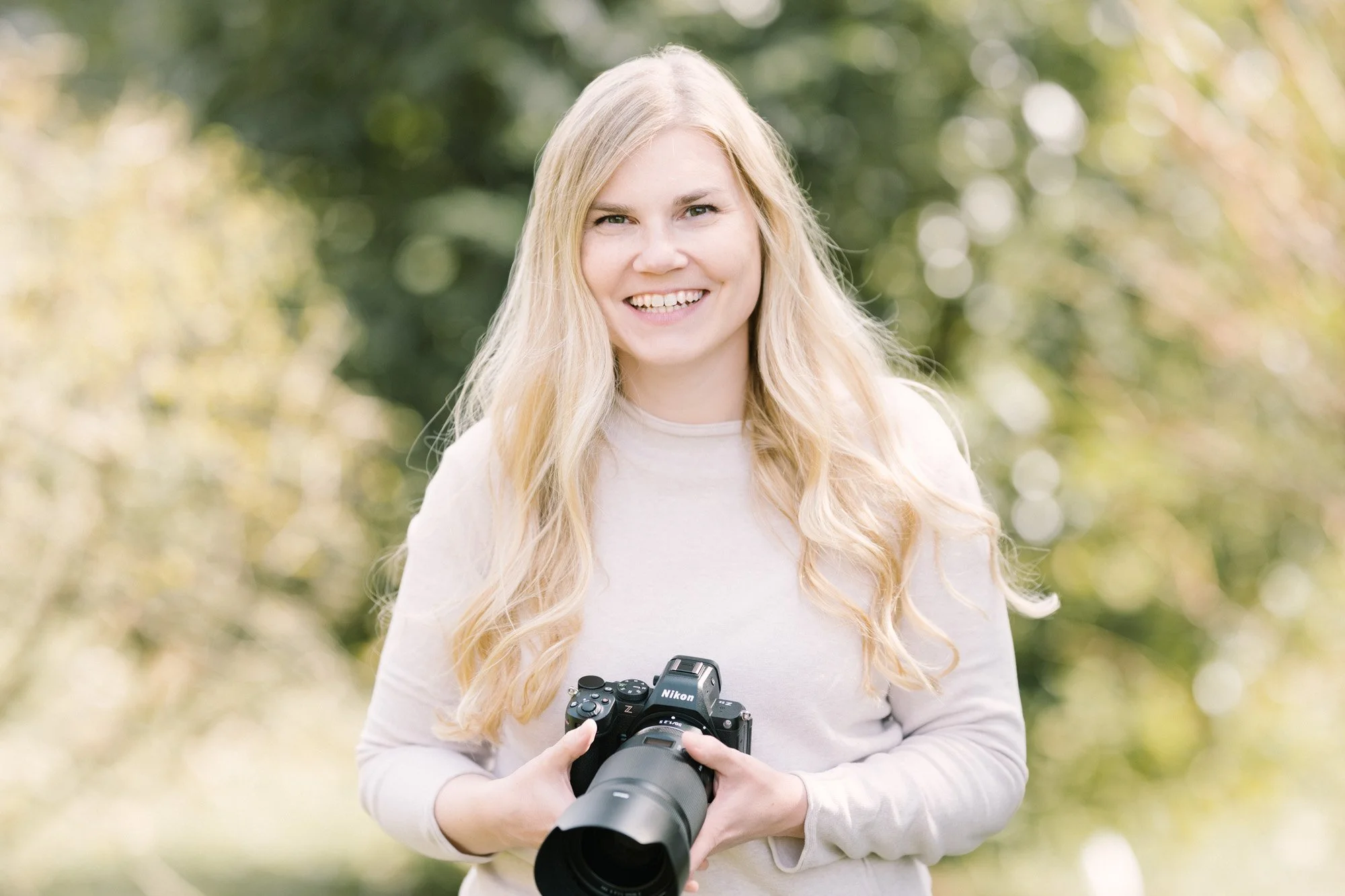 Lächelnde Frau hält eine Nikon Kamera im Freien vor einem Hintergrund aus Bäumen mit grünem Laub.