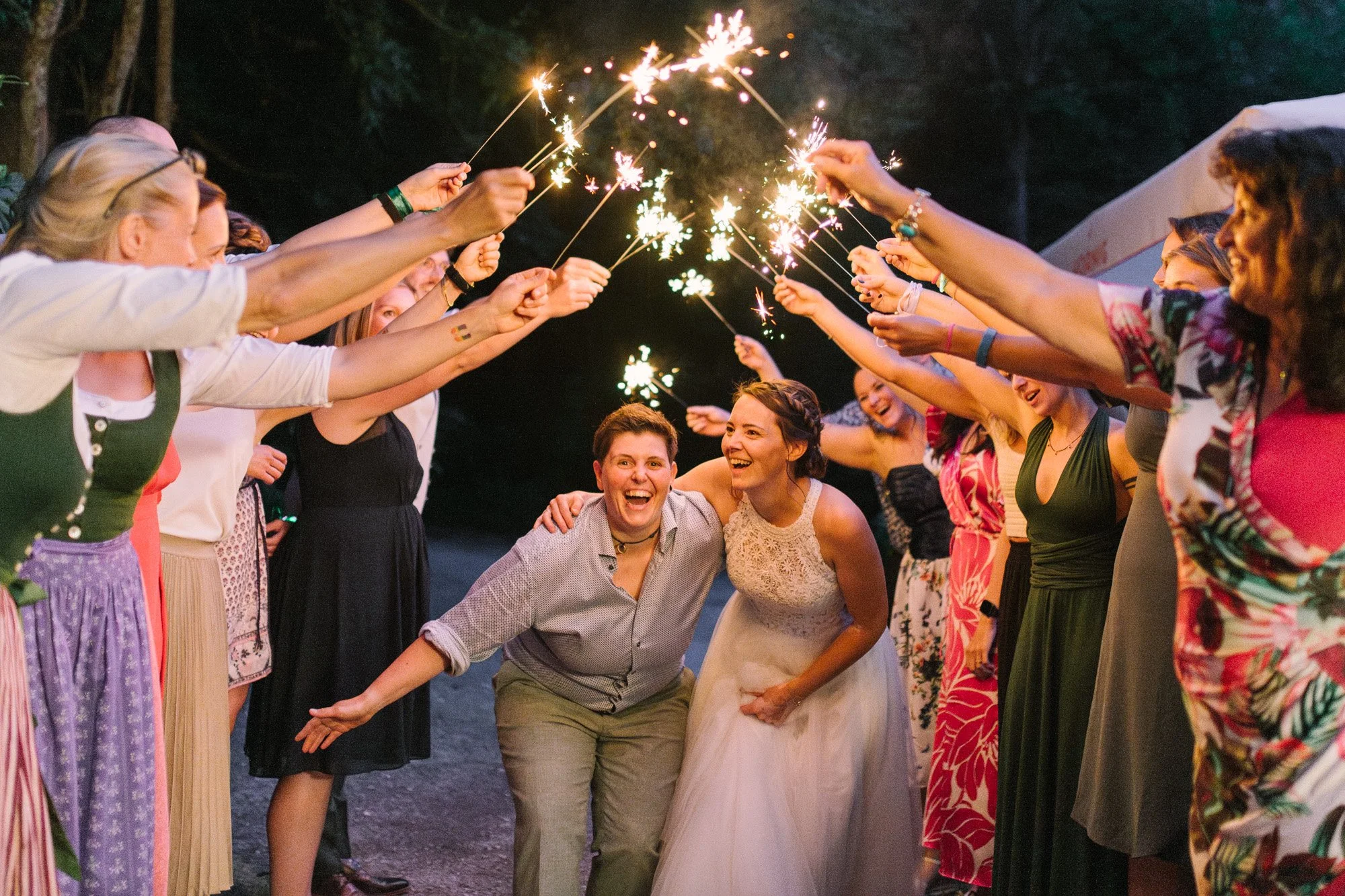 Menschen feiern bei einer Hochzeit, indem sie mit Spatzenwerkskättern Sekt in die Luft sprühen, die Braut und ein Gast lachen in der Mitte.