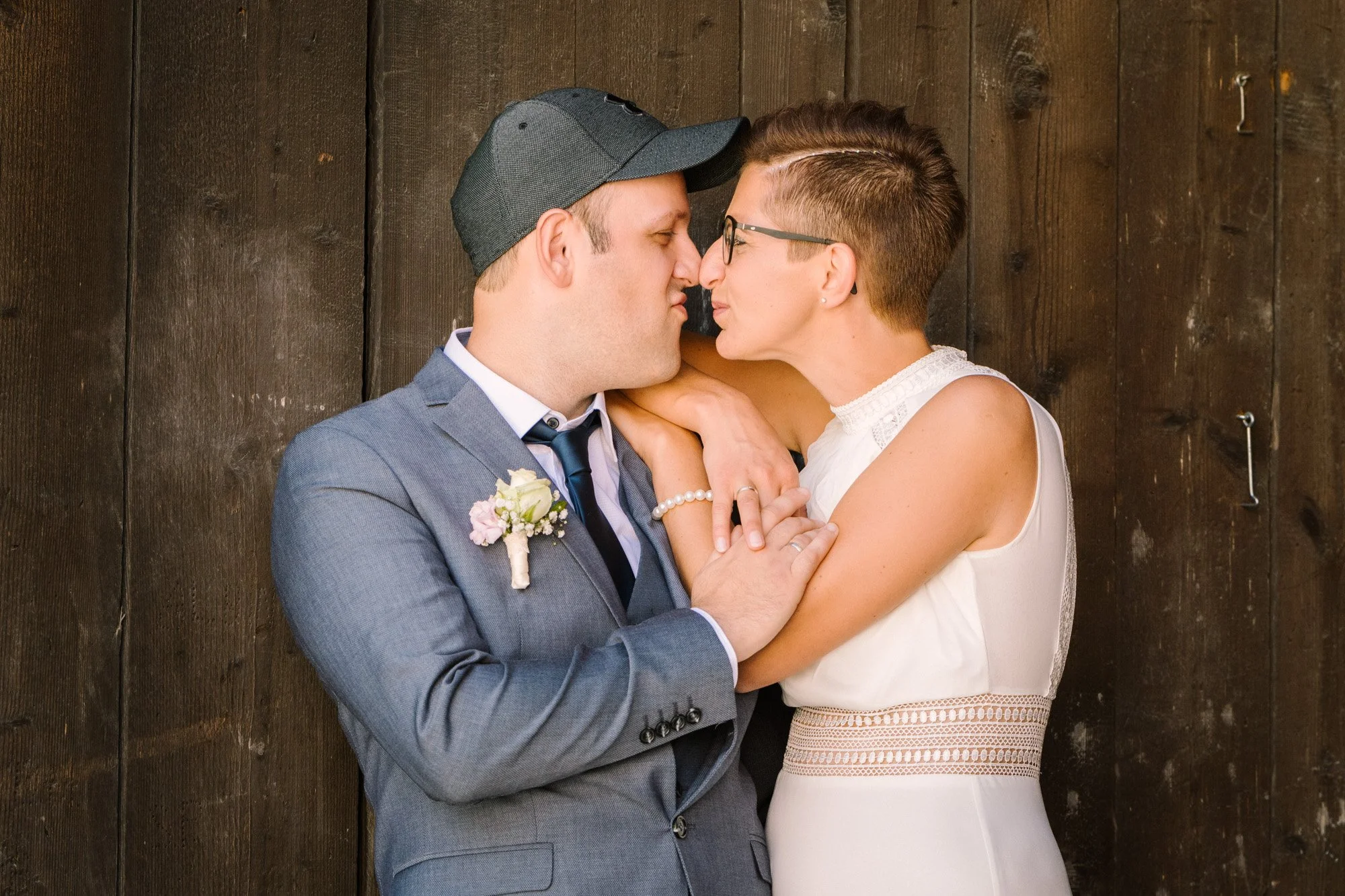 Ein Mann in Anzug und eine Frau mit kurzen Haaren und Brille stehen sich nahe vor einer Holzwand, berühren ihre Nasen und schauen sich liebevoll an.