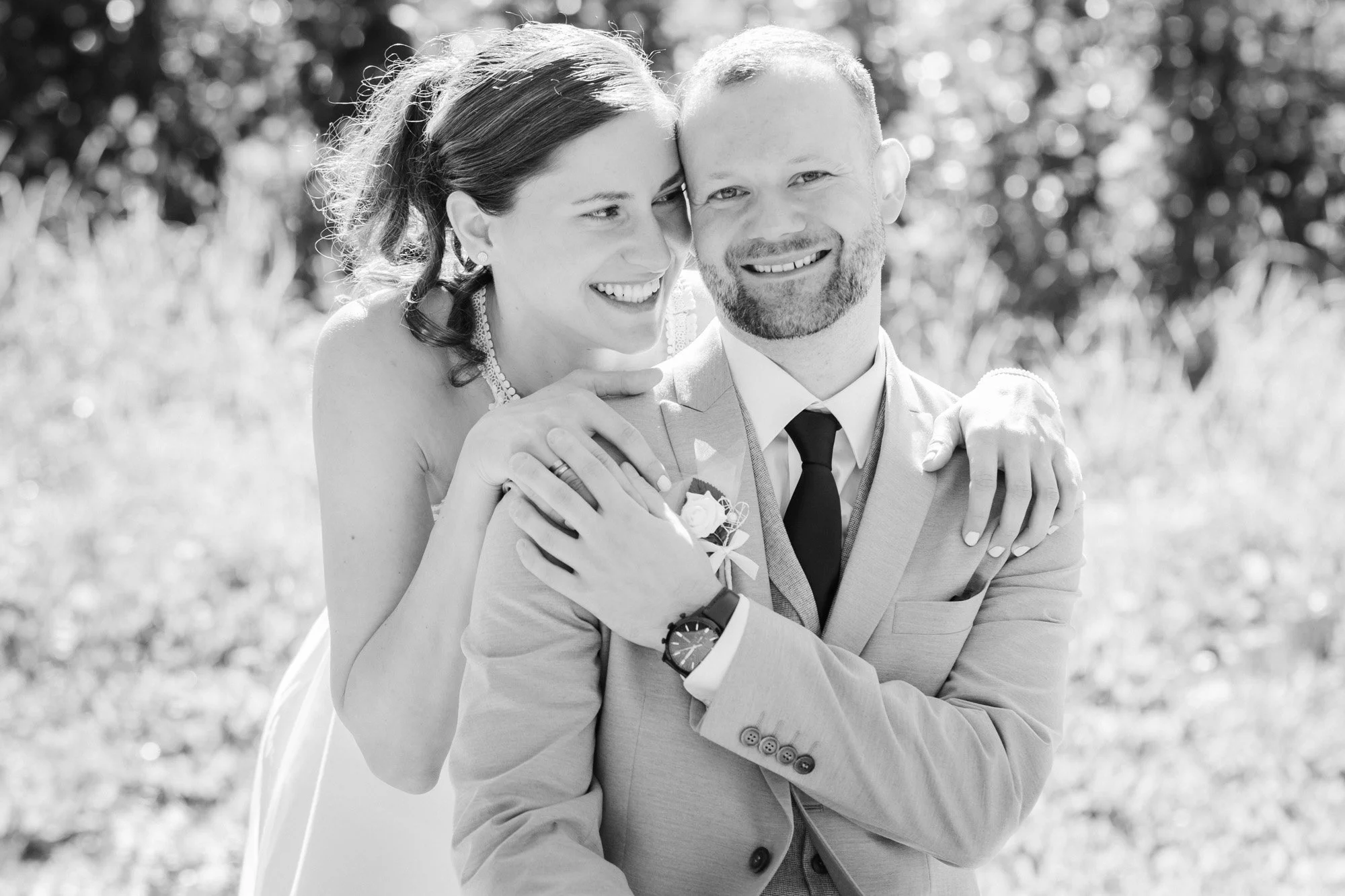 Ein glücklich wirkendes Brautpaar bei einer Hochzeit im Freien, die Frau ist liebevoll hinter dem Mann mit Armen um ihn herum, beide lächeln.