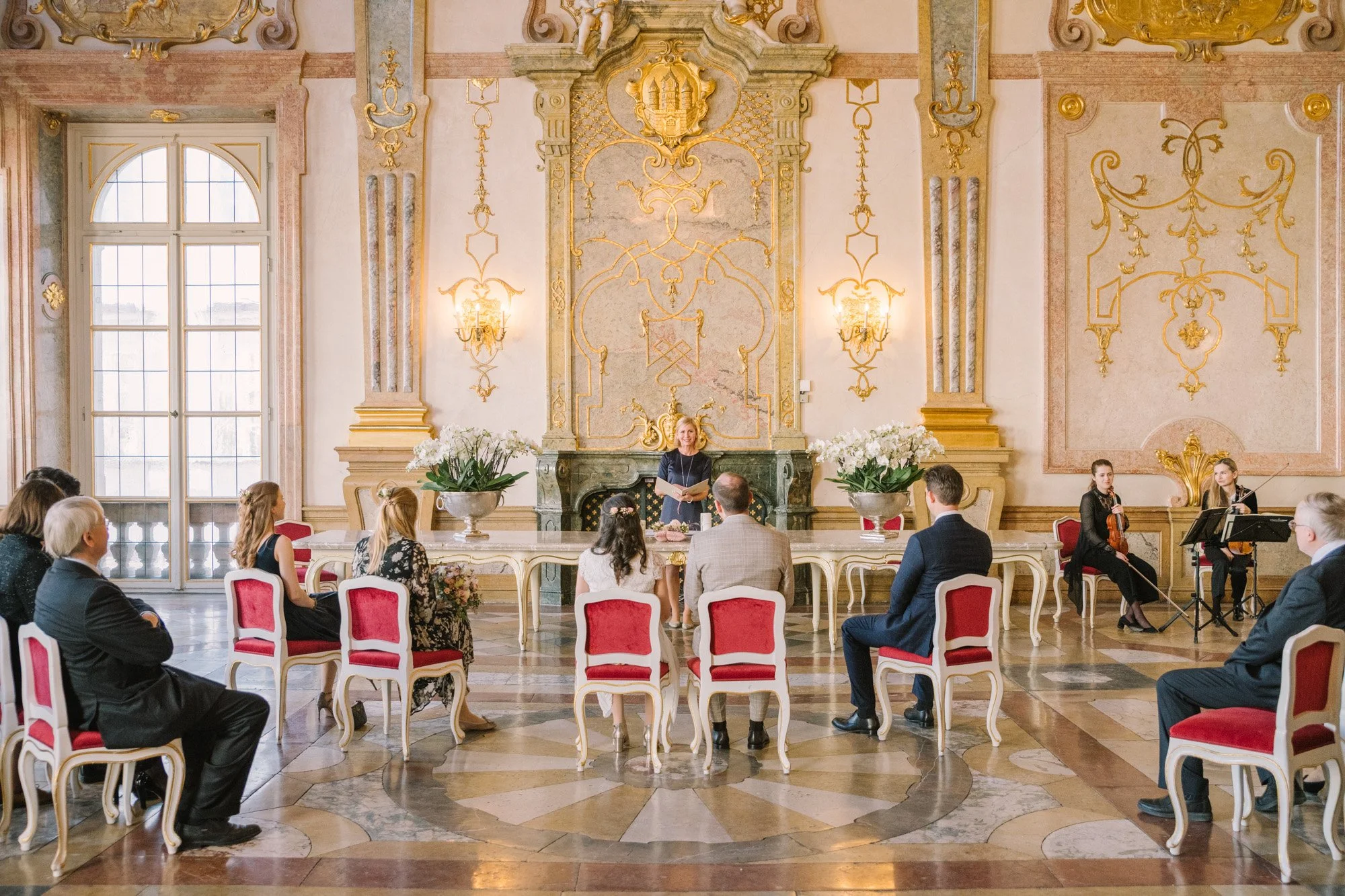 Eine formelle Veranstaltung in opulenten, goldverzierten Räumen: Eine Frau steht am Kopf eines Tisches vor einem Publikum, während zwei Musiker mit Geige auf der rechten Seite spielen, und die Zuschauer sitzen in roten Stühlen.