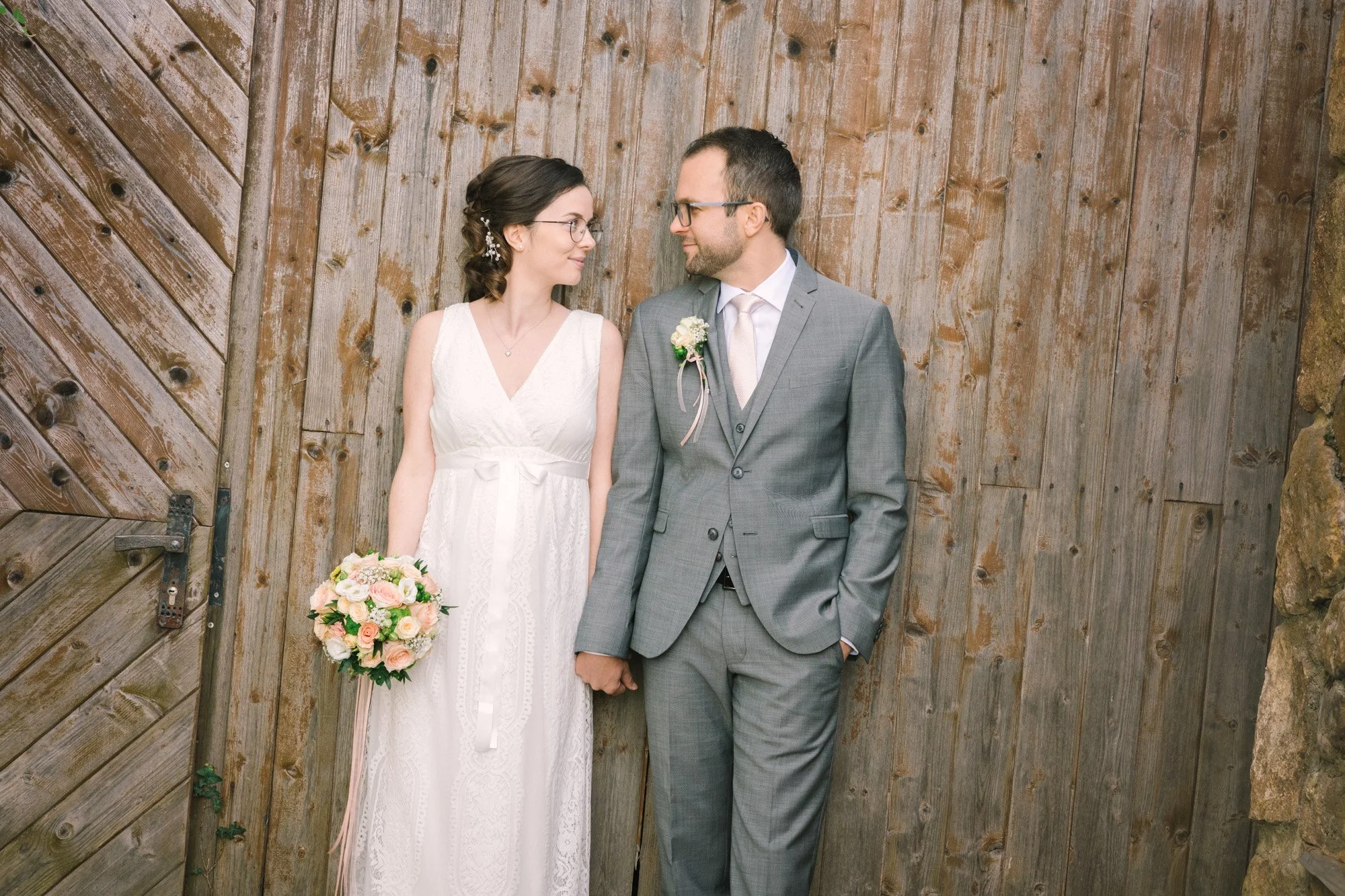 Ein Brautpaar steht vor einer Holzlatttür. Die Braut trägt ein weißes Kleid und hält einen Blumenstrauß, der Bräutigam trägt einen grauen Anzug.