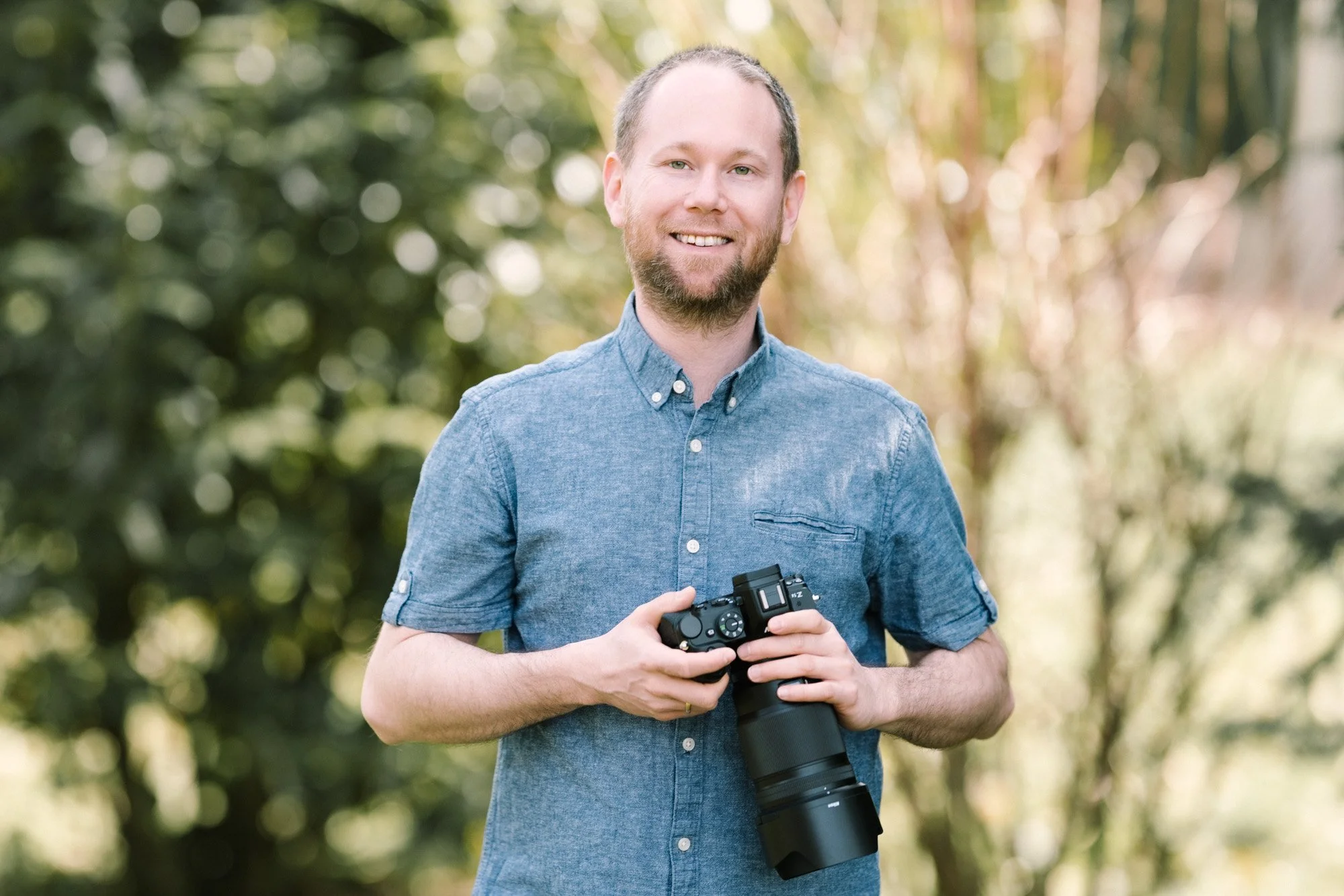 Ein lächelnder Mann mit einem blauen Hemd hält eine Kamera mit einem langen Teleobjektiv vor einem bewaldeten Hintergrund.