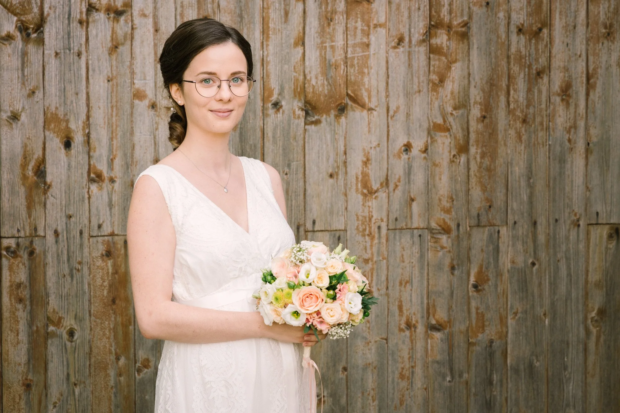 Frau in weißem Brautkleid hält einen Blumenstrauß, steht vor einer Holzwand.