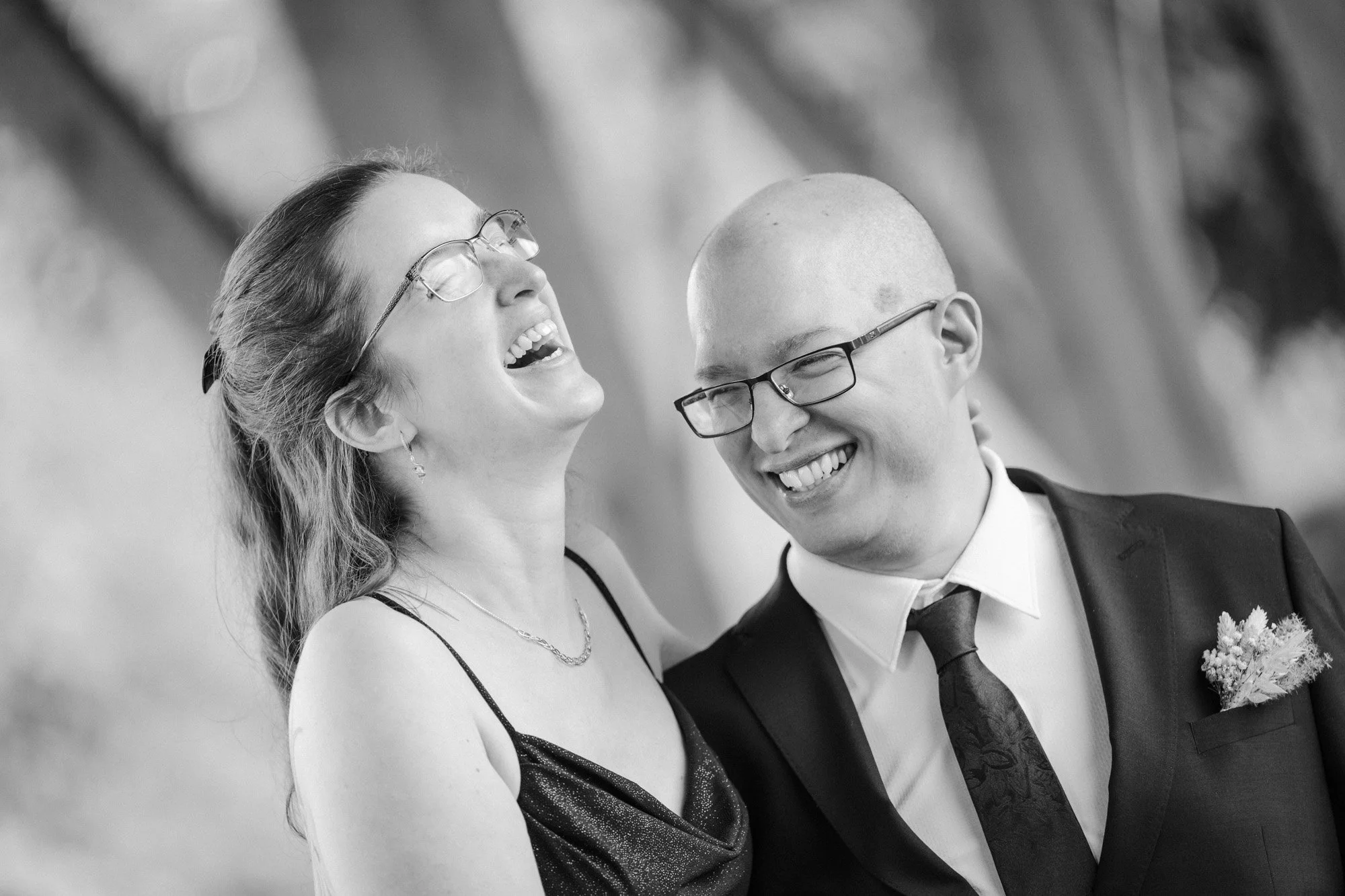 Zwei Menschen lachen fröhlich bei einer formellen Gelegenheit, eine Frau mit langen Haaren und Brille, ein Mann mit Glatze und Brille, im Anzug mit Krawatte und Blume im Revers.