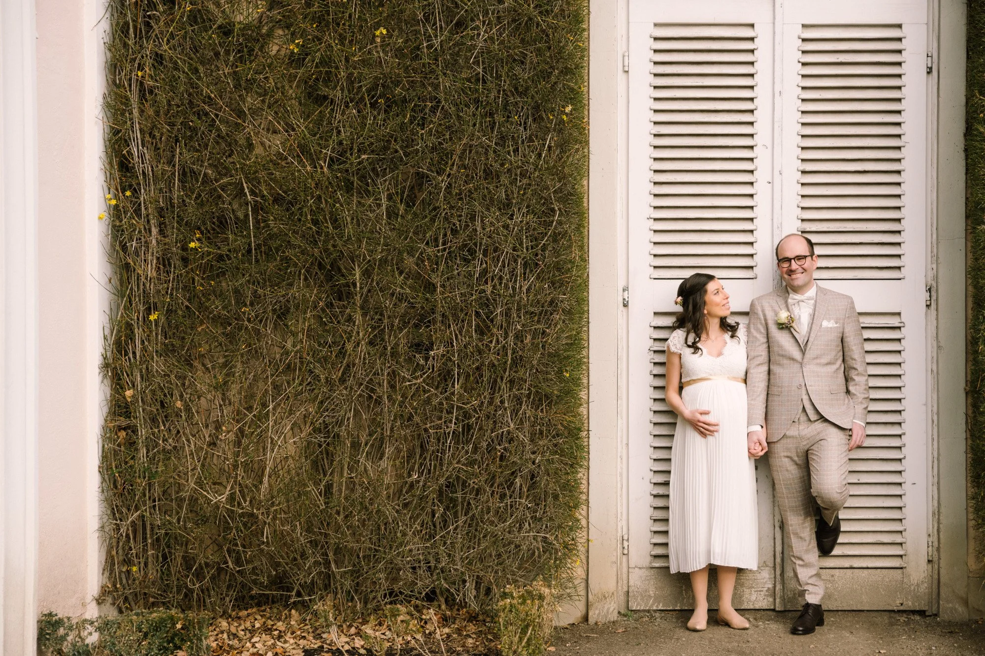 Ein braun gekleideter Mann im Anzug mit Brille und ein schwangere Frau in einem weißen Kleid, die sich an den Händen halten, lehnen an einer weißen Tür mit geschlitzten Lamellen, während sie sich ansehen.