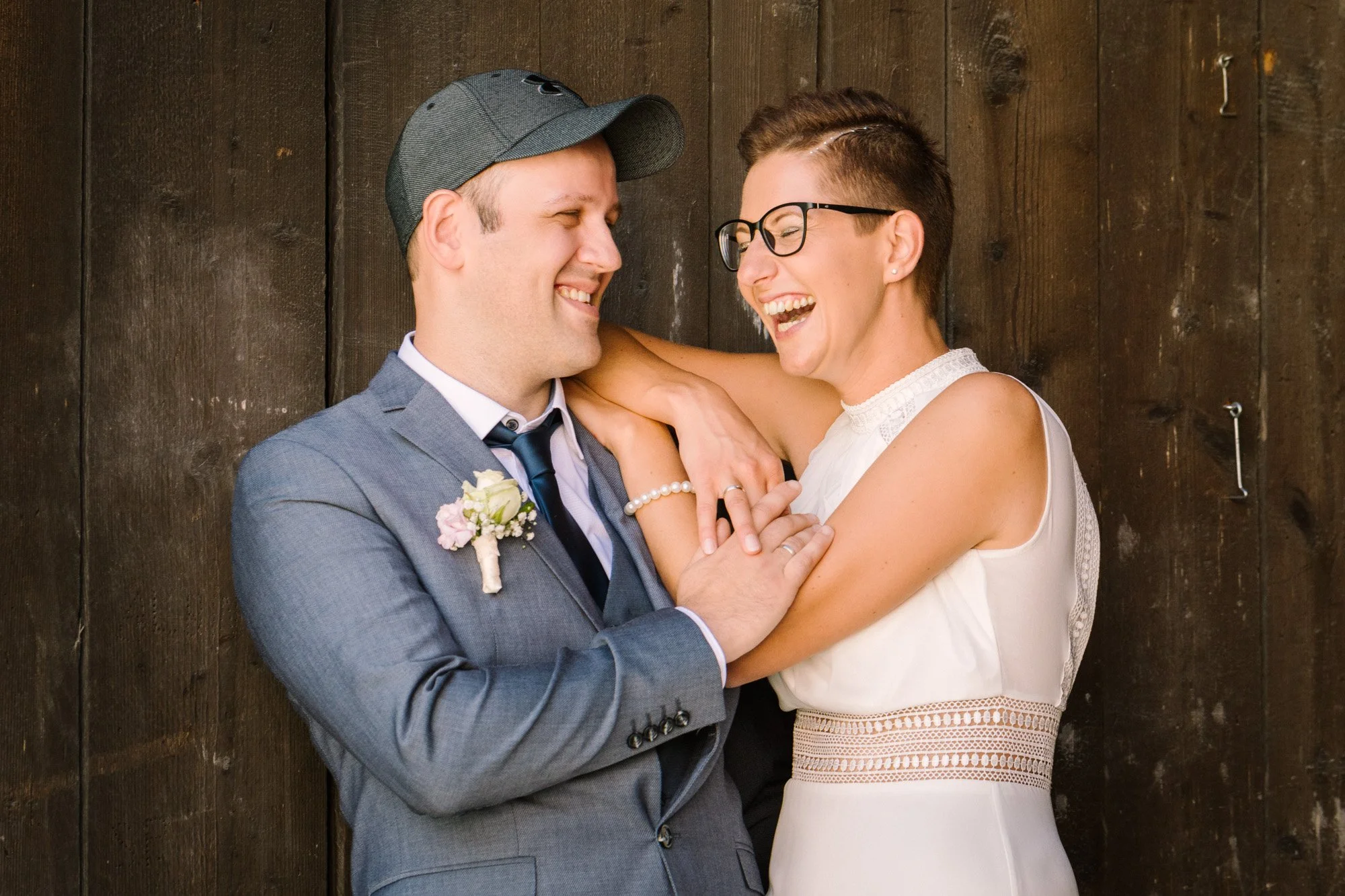 Ein lachendes Paar bei einer Hochzeit, umgeben von einer dunklen Holzwand, in formeller Kleidung, die sich liebevoll umarmen.