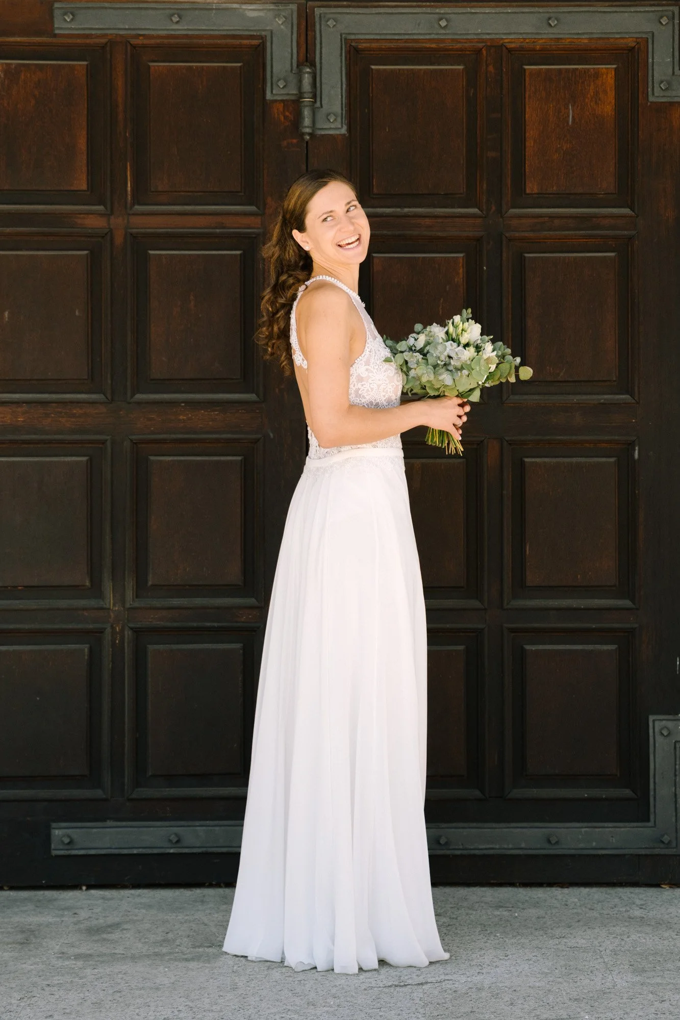 Braut in weißem Kleid mit Blumenstrauß vor einer dunklen Holzgarage.