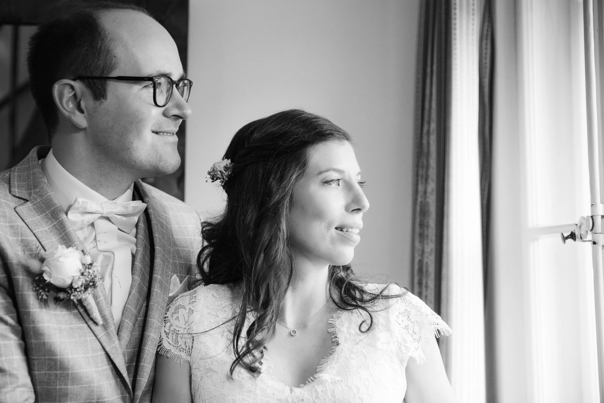 Ein Mann und eine Frau schauen durch ein Fenster und lächeln. Der Mann trägt eine Brille, einen Anzug mit Krawatte, die Frau trägt ein weißes Kleid mit Spitze und hat Haare mit Blumen geschmückt.