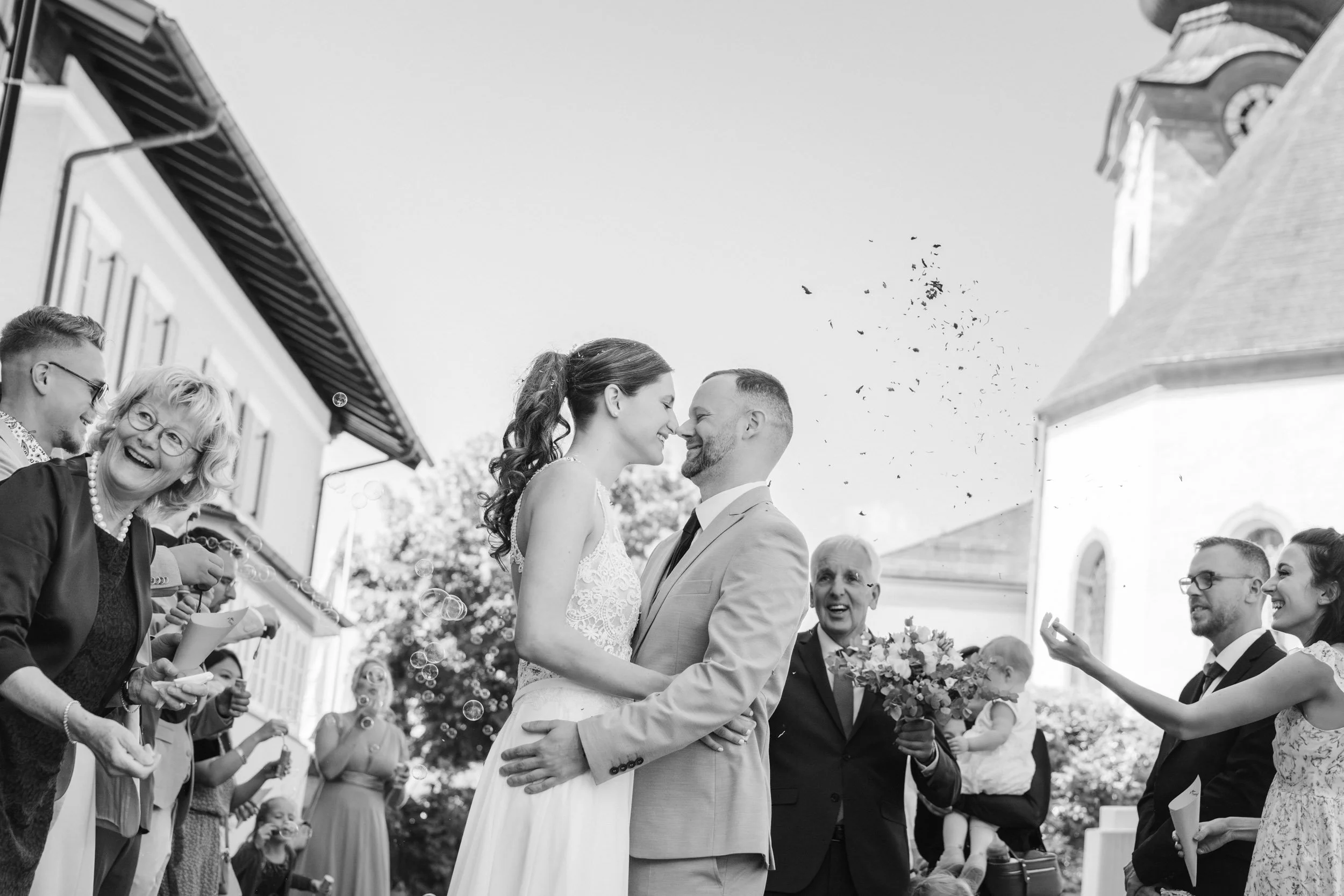 Hochzeitspaare stehen sich gegenüber, umgeben von Freunden und Familienmitgliedern, die feiern, mit einem älteren Mann, der einen Blumenstrauß hält, im Hintergrund. Es scheint eine festliche, glückliche Szene zu sein.