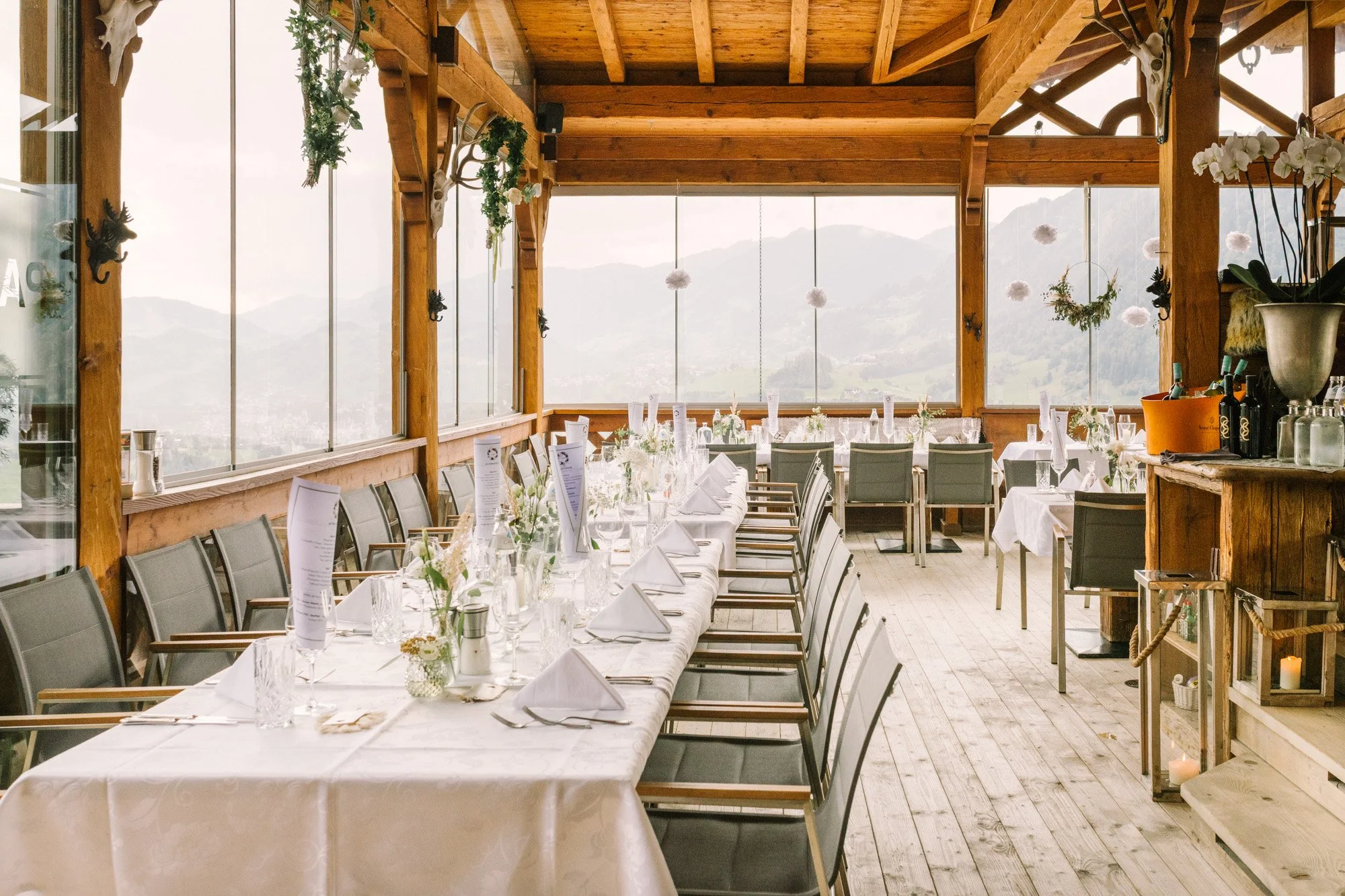 Ein festlich gedeckter Speisesaal mit langen weißen Tischen, weißen Servietten, Gläsern und kleinen Blumenarrangements, in einem rustikalen Raum mit großen Fenstern, Blick auf Berge, und Dekorationen aus weißen Blumen und Kerzen.