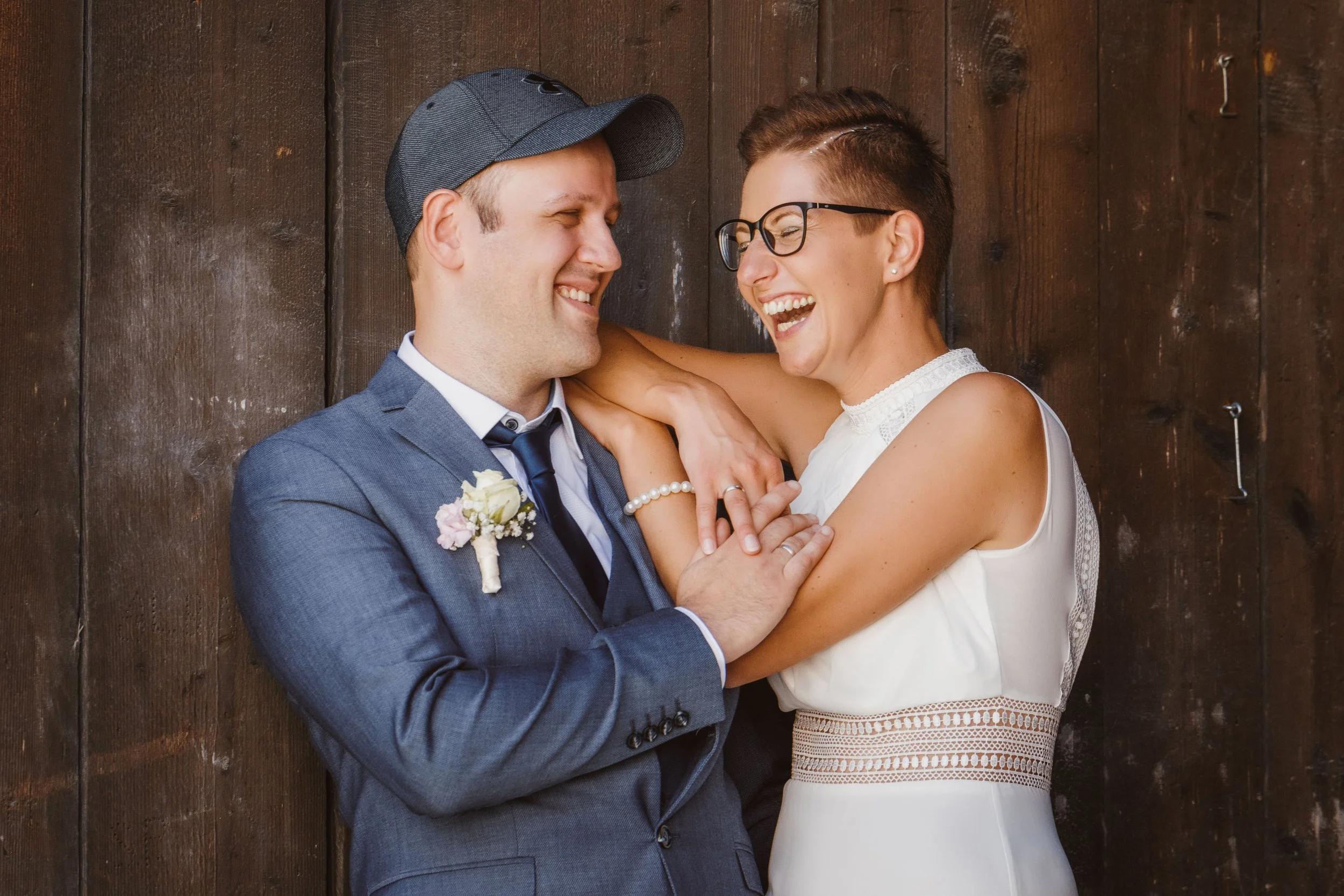 Ein lächelndes Paar bei einer Hochzeit, die Frau trägt ein weißes Kleid, der Mann einen grauen Anzug und eine Kappe, sie umarmen sich vor einer dunklen Holzvertäfelung.