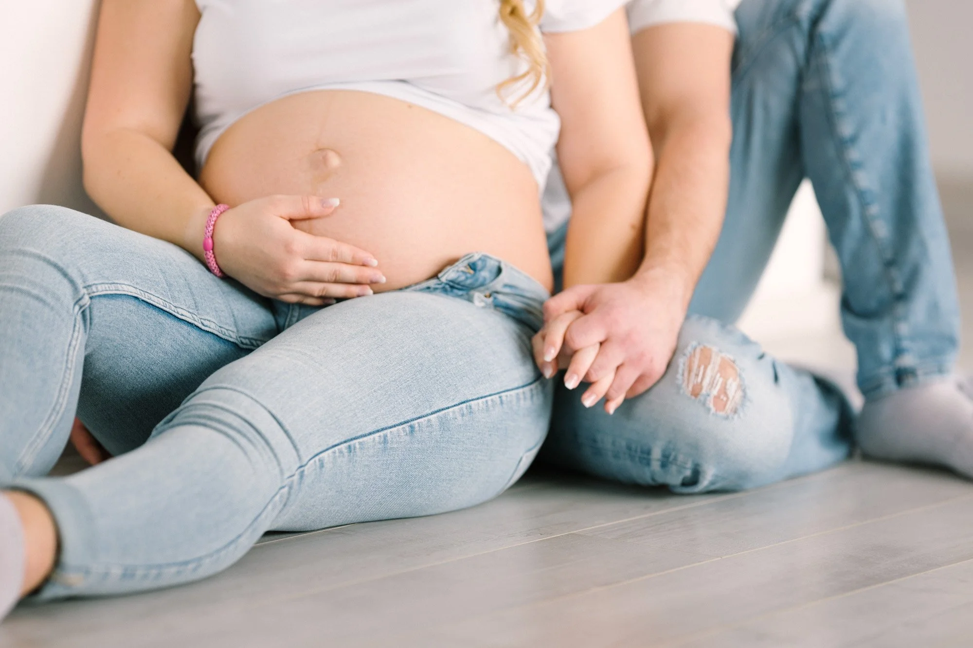 Schwangere Frau and Partner sitzen auf dem Boden, halten Hände auf den Bauch, tragen Jeans. Nahaufnahme, entspannt, liebevoll.