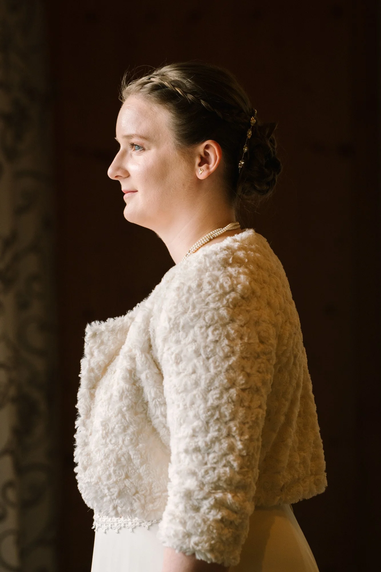 Eine Frau in Hochzeitskleidung, im Profil gesehen, mit eleganter Hochsteckfrisur und Perlenkette.