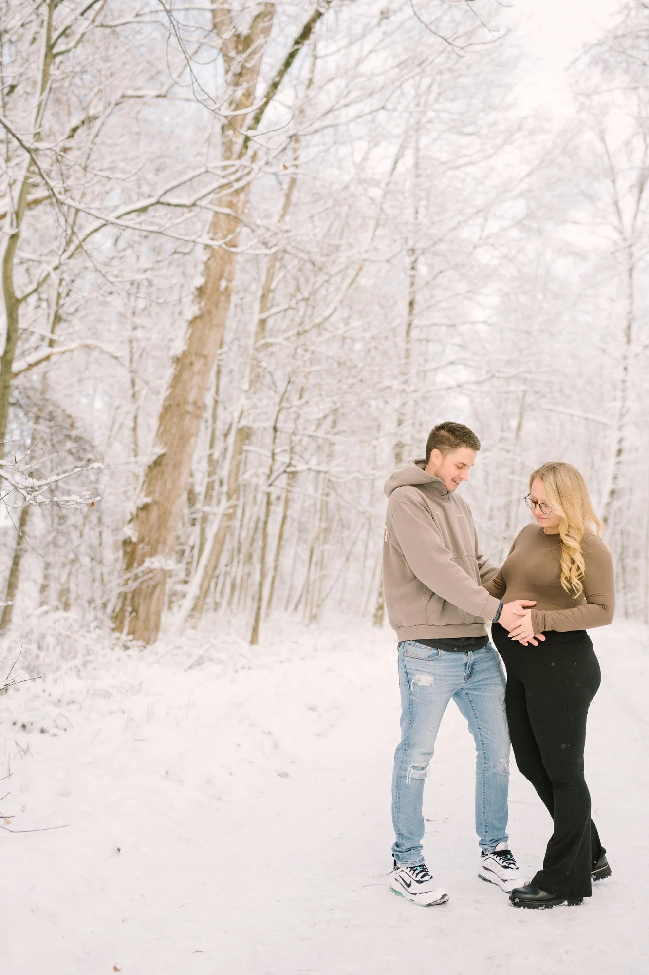 Ein Paar steht im Schnee im Winterwald, die Frau ist schwanger, sie hält ihren Bauch, während der Mann sie liebevoll berührt.