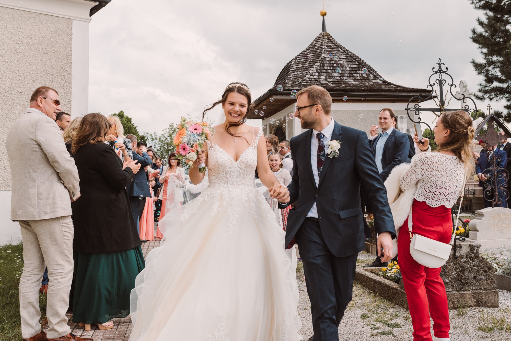 Braut und Bräutigam gehen bei ihrer Hochzeit im Freien Hand in Hand, umgeben von lachenden Gästen. Die Braut trägt ein weißes Hochzeitskleid mit Blumenstrauß, der Bräutigam trägt einen dunklen Anzug. Es gibt Seifenblasen im Himmel und eine historische Kapelle im Hintergrund.