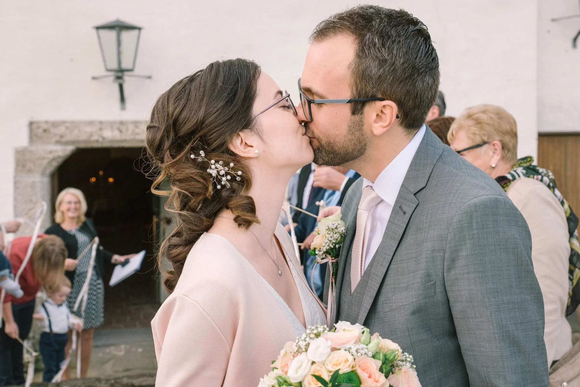 Ein Brautpaar küsst sich bei einer Hochzeit, im Hintergrund feiern Gäste in einer alten Steinhalle.