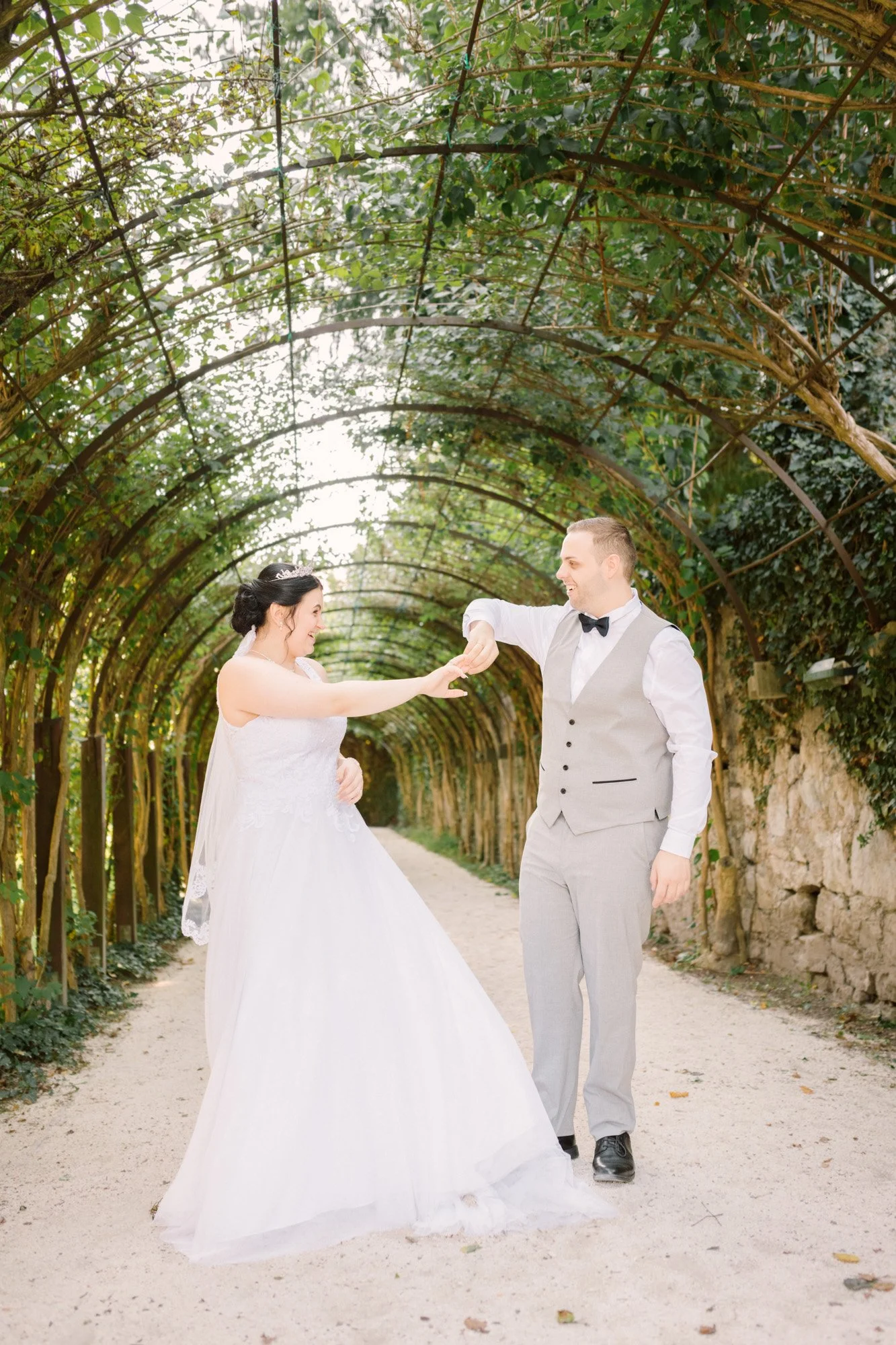 Ein Brautpaar tanzt unter einer gewölbten Laube aus grünen Blättern auf einem Gartenweg.
