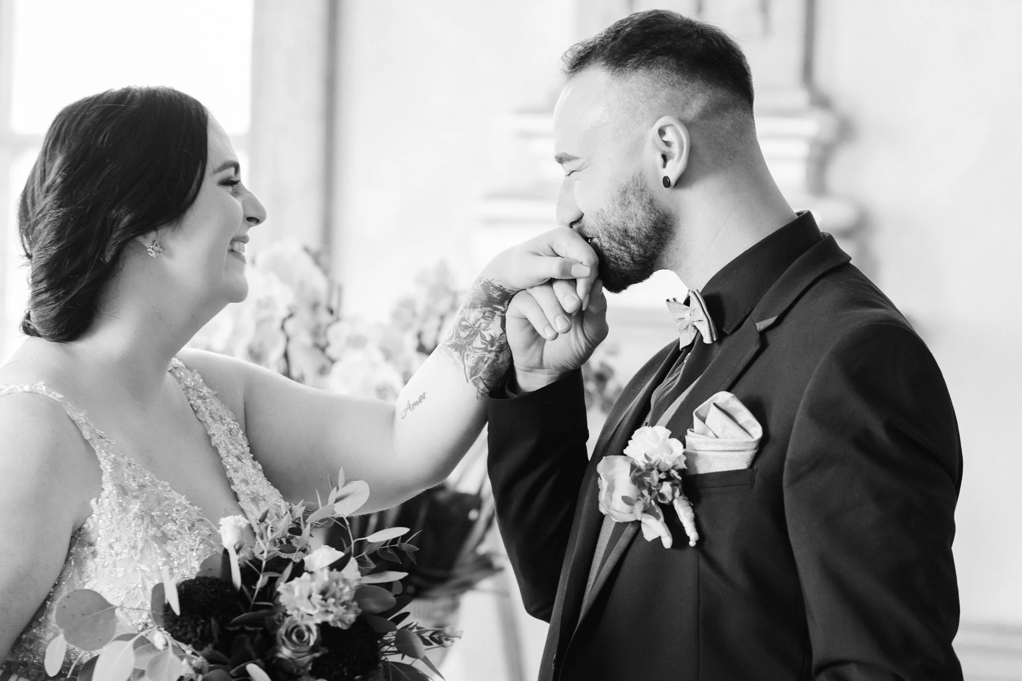 Ein Mann auf einer Hochzeit küsst zärtlich die Hand einer lachenden Frau mit Brautkleid, im Hintergrund Blumen und ein Fenster.