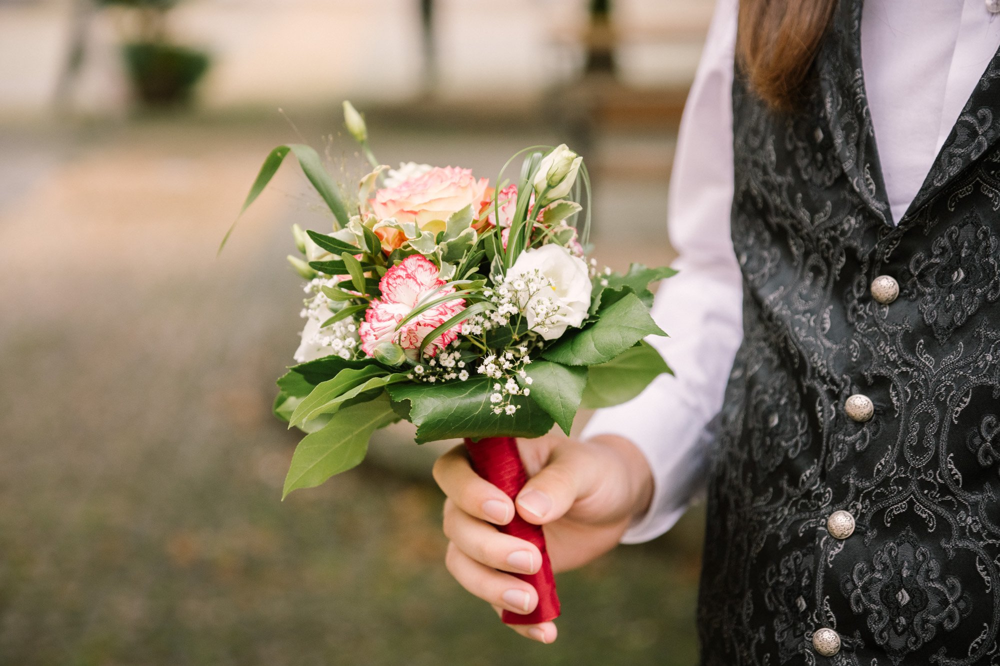 Jemand hält einen kleinen Blumenstrauß mit verschiedenen Blumen und grünen Blättern, der mit rotem Stoff umwickelt ist.