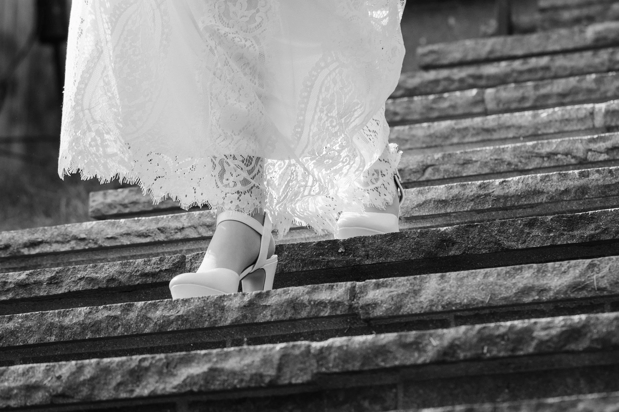 Nahaufnahme einer Person, die auf einer Steintreppe in einem langen, weißen, lace-geschmückten Kleid mit hohen, eleganten Schuhen steht.