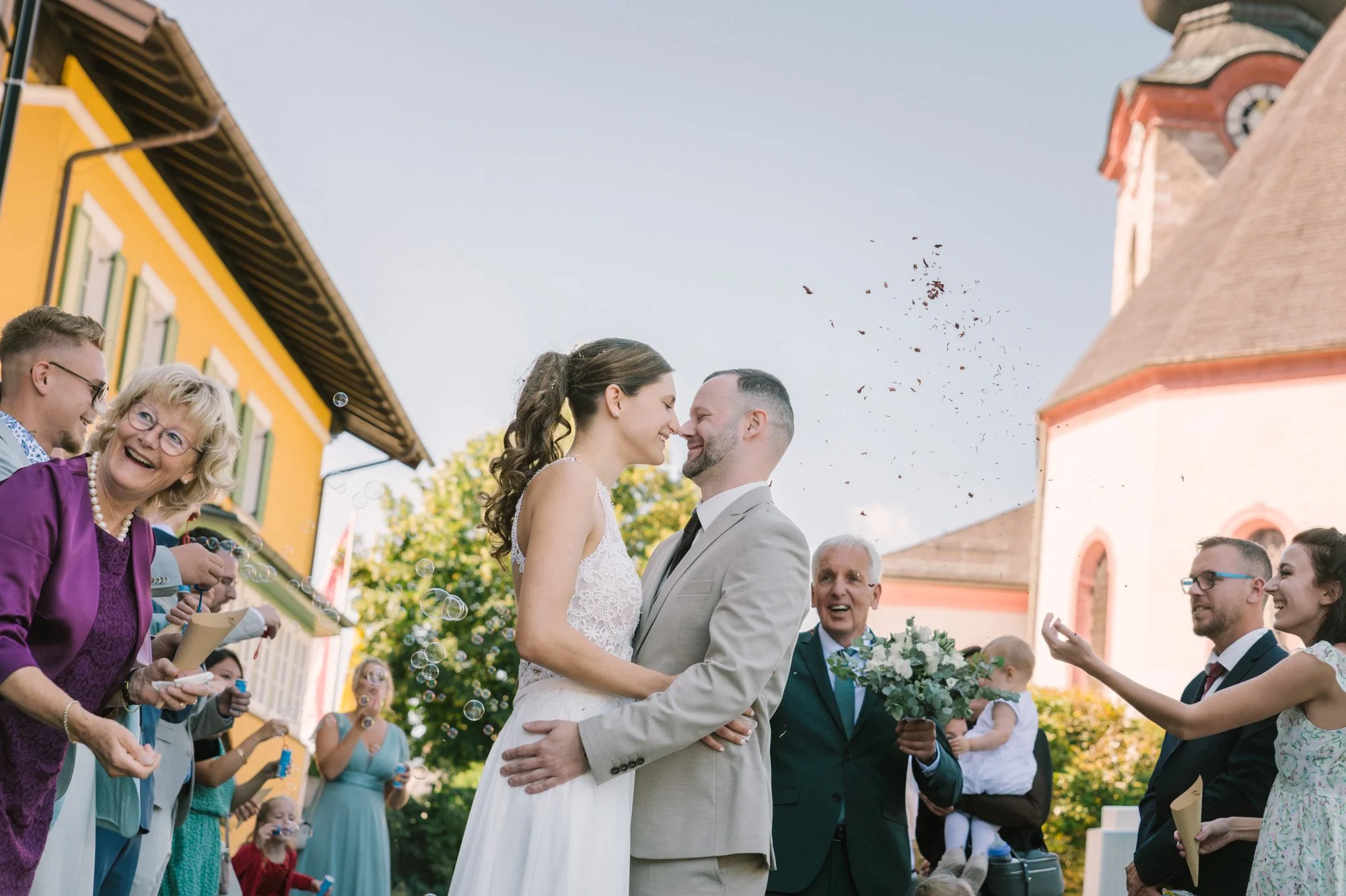 Ein Brautpaar steht sich bei einer Hochzeitsfeier im Freien lachend gegenüber, während Gäste um sie herum fröhlich feiern und Blasen pusten. Im Hintergrund sind bunte Gebäude und eine Kirche zu sehen.