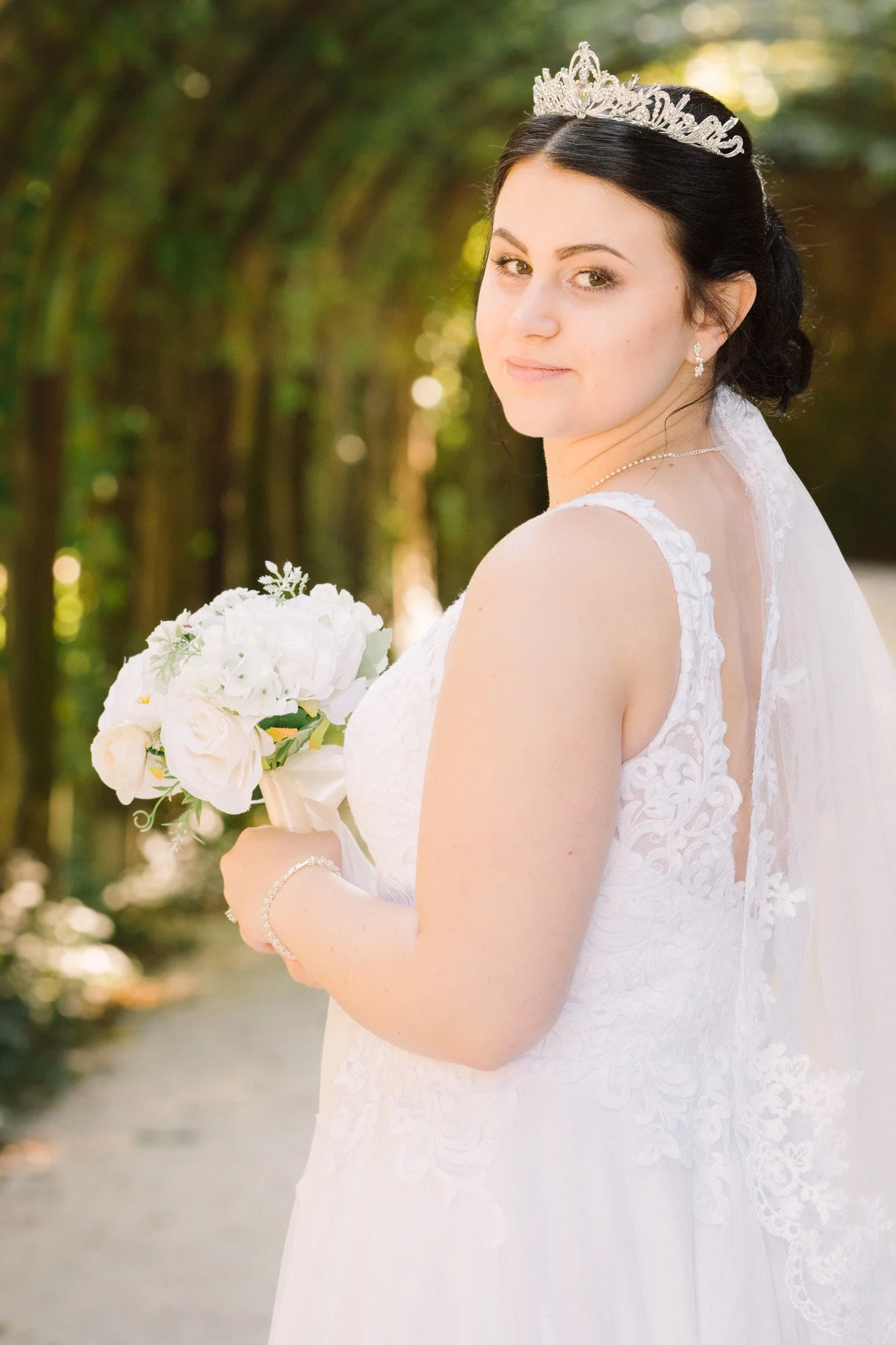 Braut in weißem Hochzeitskleid mit Diadem und Schleier, hält einen Blumenstrauß, steht im Freien vor einer Baumreihe, schaut zur Kamera.