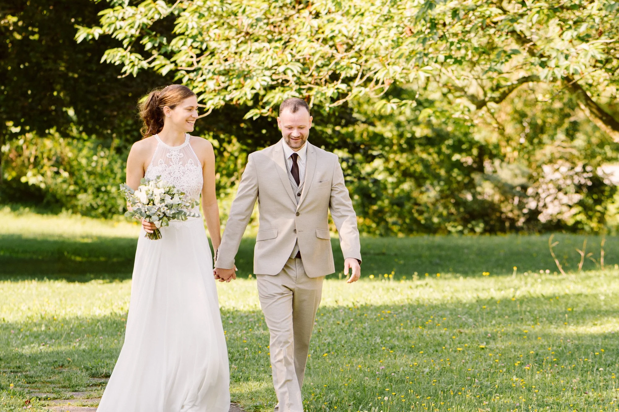 Ein frisch verheiratetes Paar geht Hand in Hand durch einen Park an einem sonnigen Tag, die Frau trägt ein weißes Brautkleid und hält einen Blumenstrauß, der Mann trägt einen grauen Anzug und eine Krawatte.