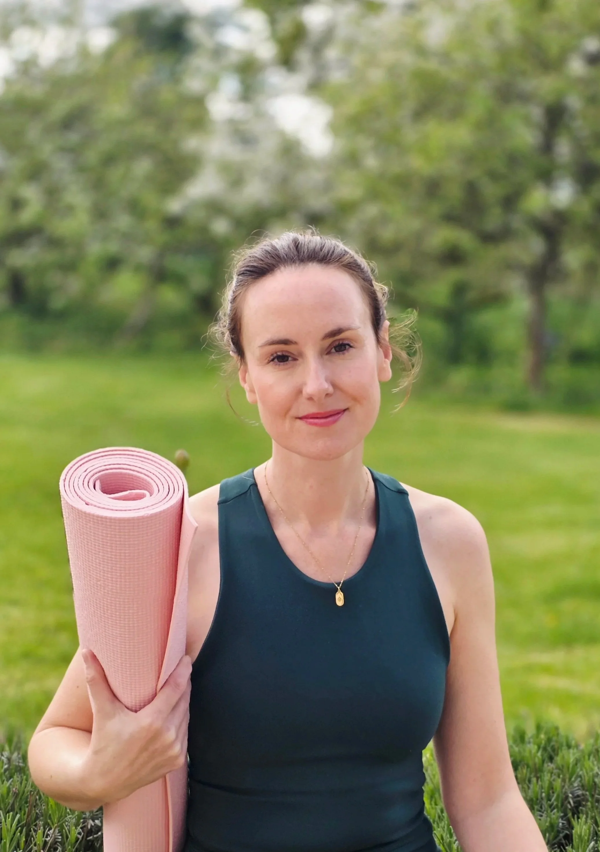A woman in a black sleeveless top holding a pink yoga mat on her shoulder, outdoors in a grassy area with trees in the background.